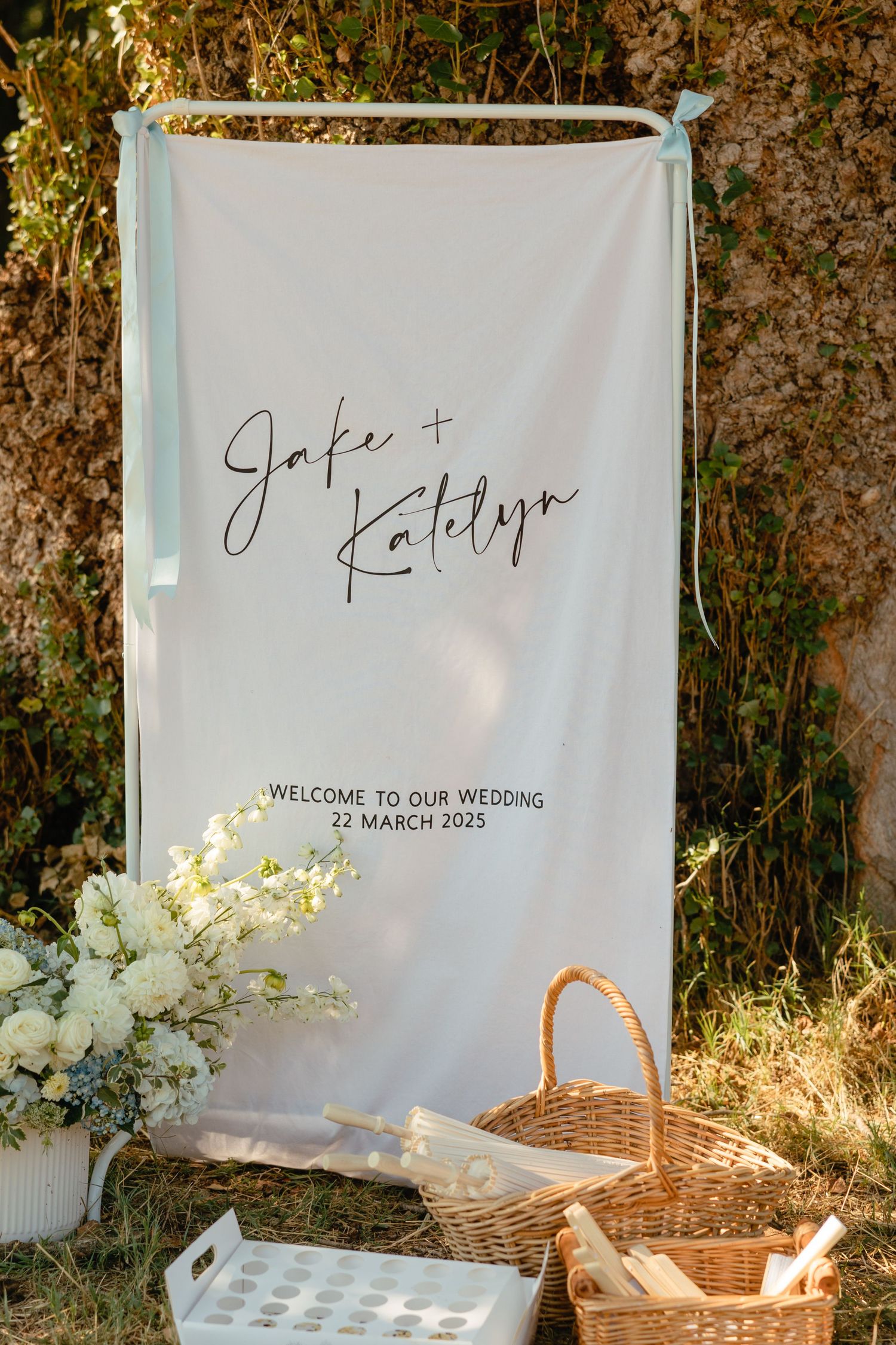 Elegant white wedding welcome banner with the couple's names displayed above wicker baskets and floral arrangements against foliage.