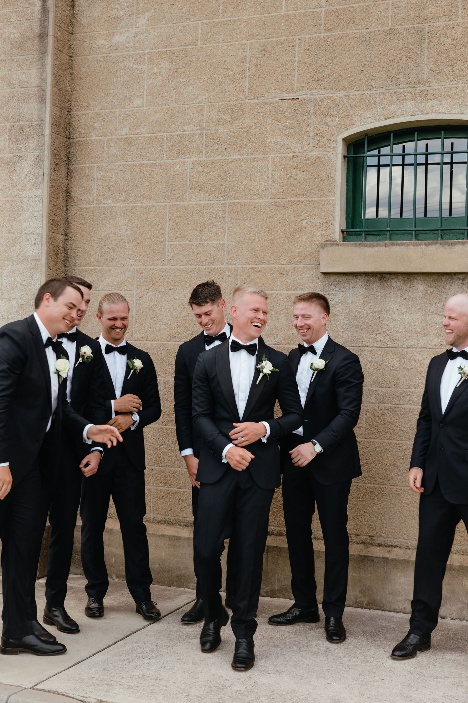 A group of groomsmen in black tuxedos share a candid laughing moment against a stone wall at a wedding.