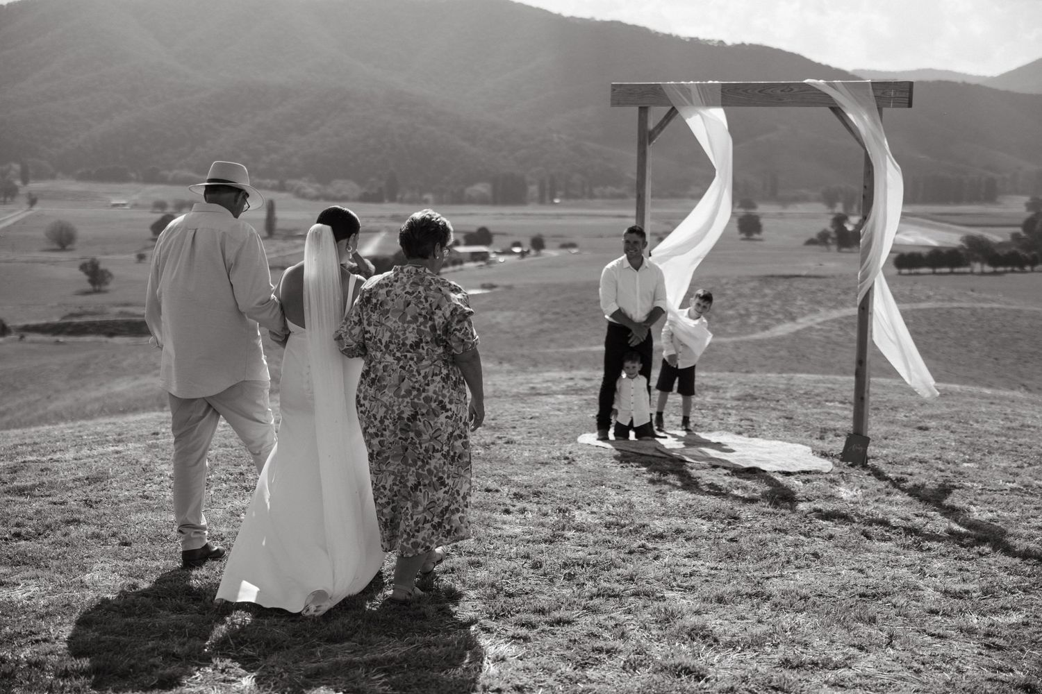 Wedding ceremony taking place on a hilltop with white flowing fabric arch and rolling mountains in the background.