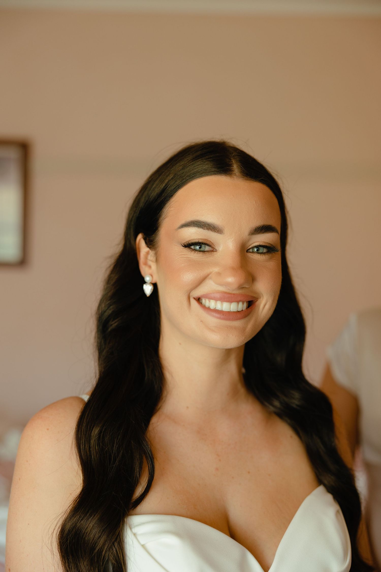 A bride with long dark hair and pearl earrings smiles radiantly in natural lighting.