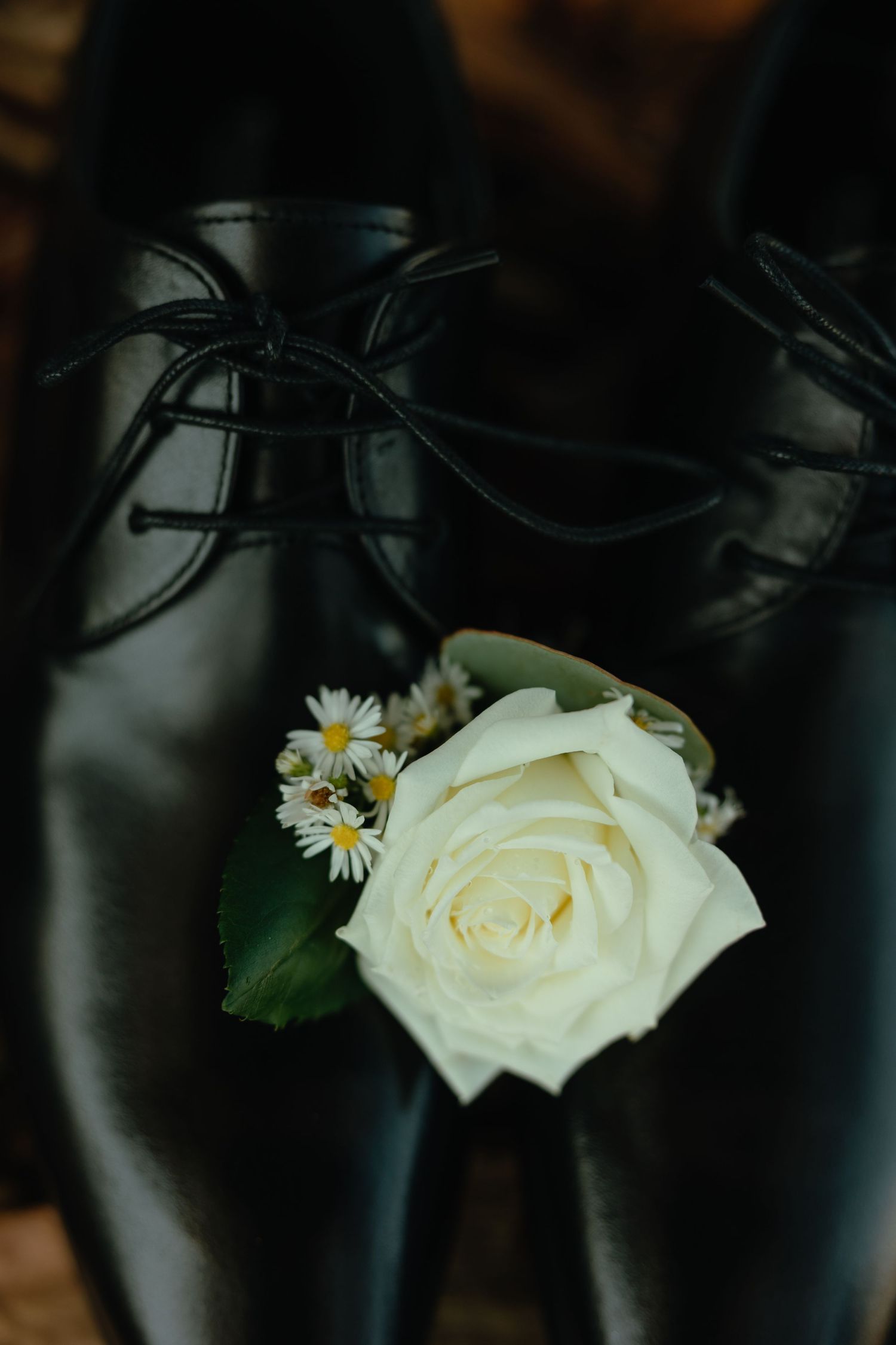 A white rose and daisy boutonniere rests atop black formal leather shoes.