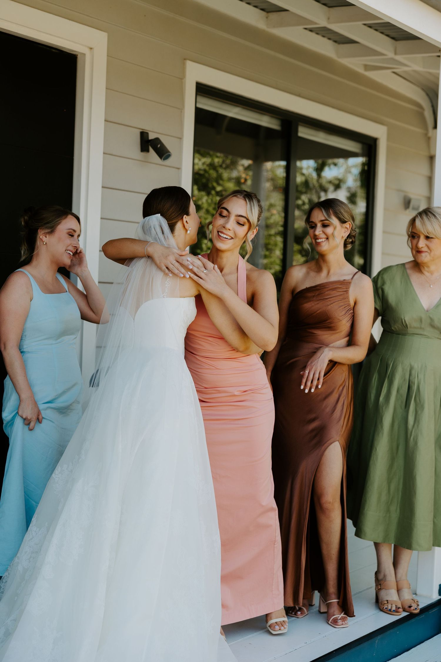 Wedding party shares candid laughter together on a covered porch wearing colorful bridesmaid dresses.
