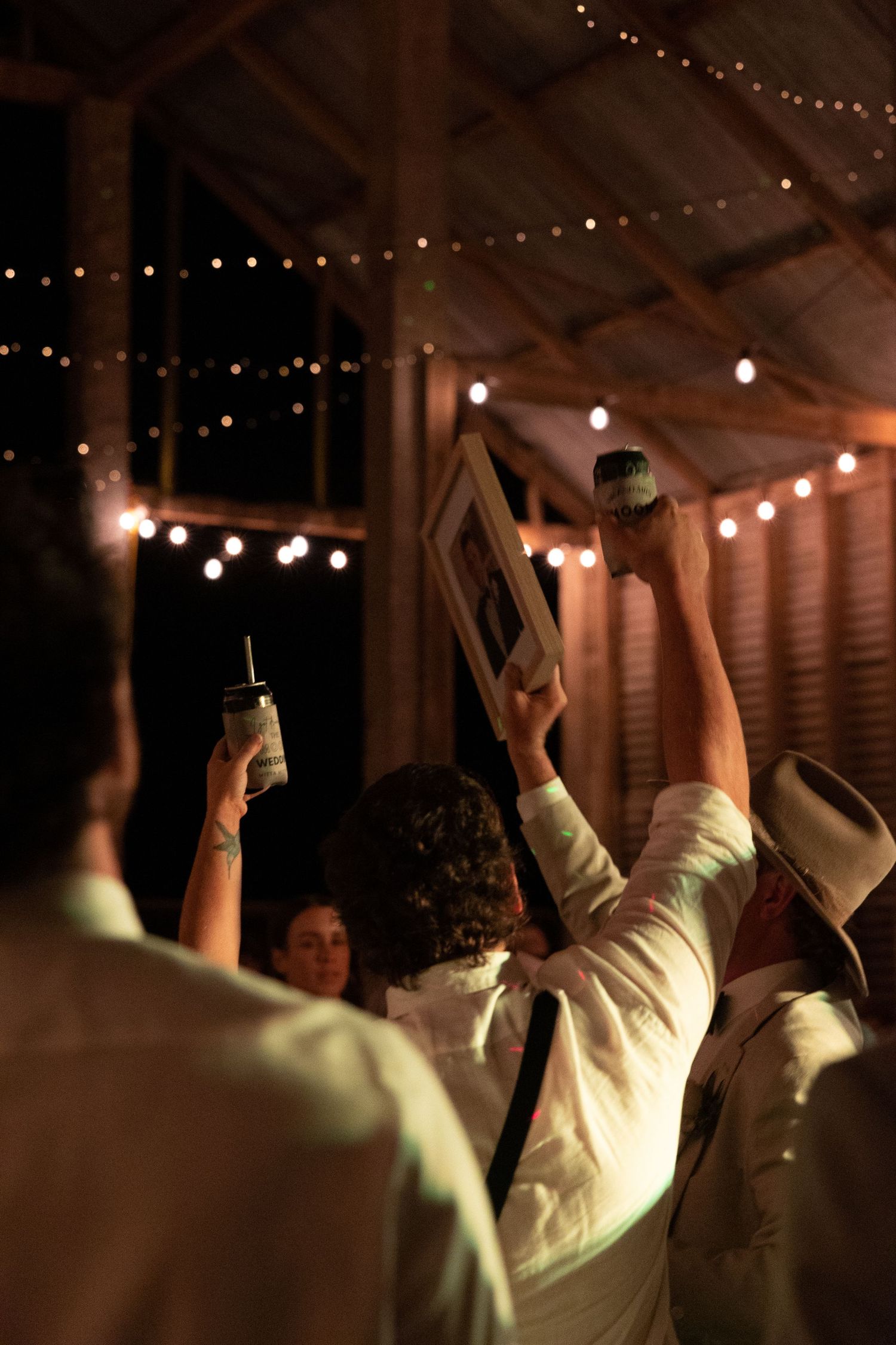 People dancing and celebrating at a rustic indoor venue decorated with string lights overhead during a party or reception.