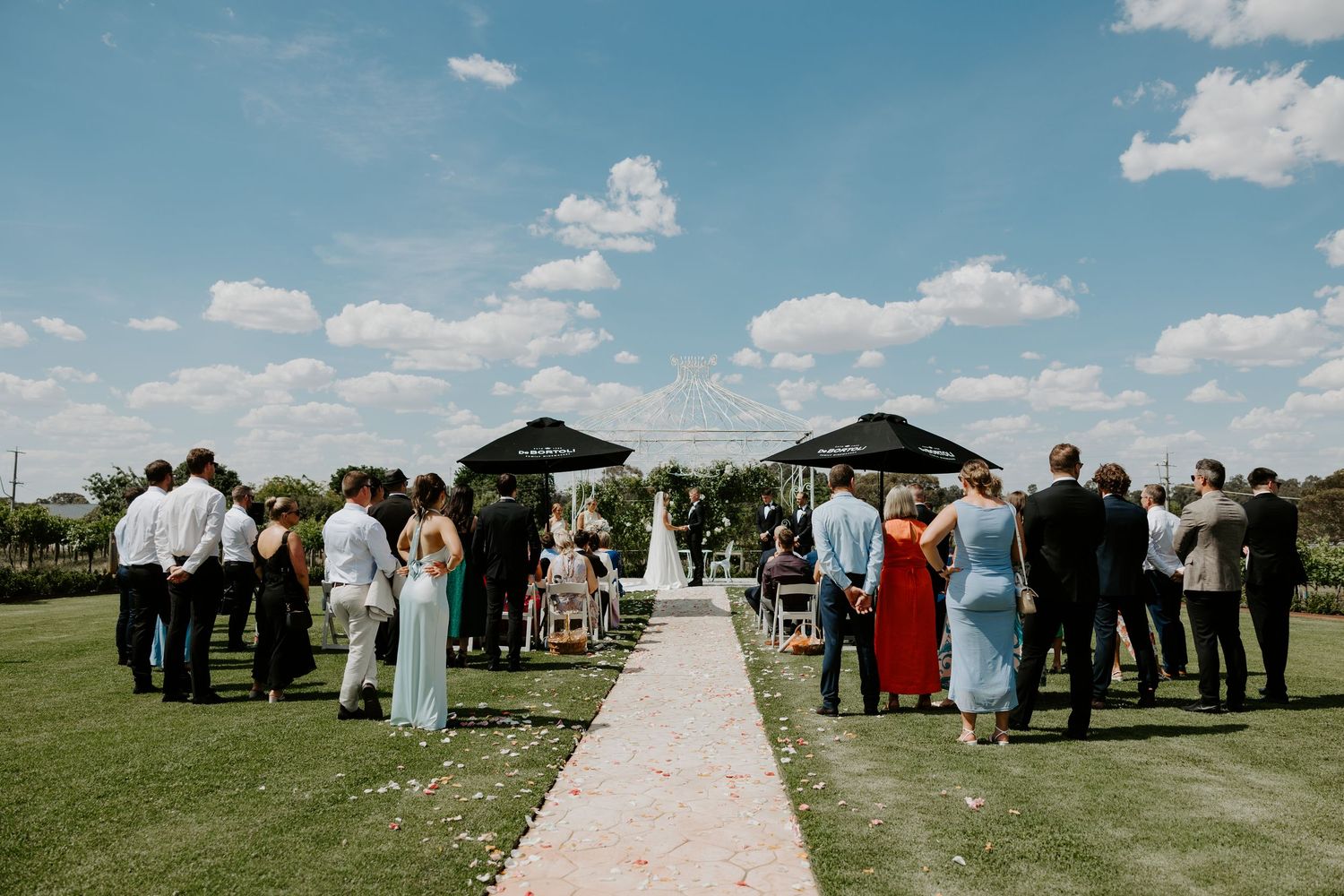 Wedding guests gather on a lush green lawn alongside a white petal-lined aisle path under blue summer skies.