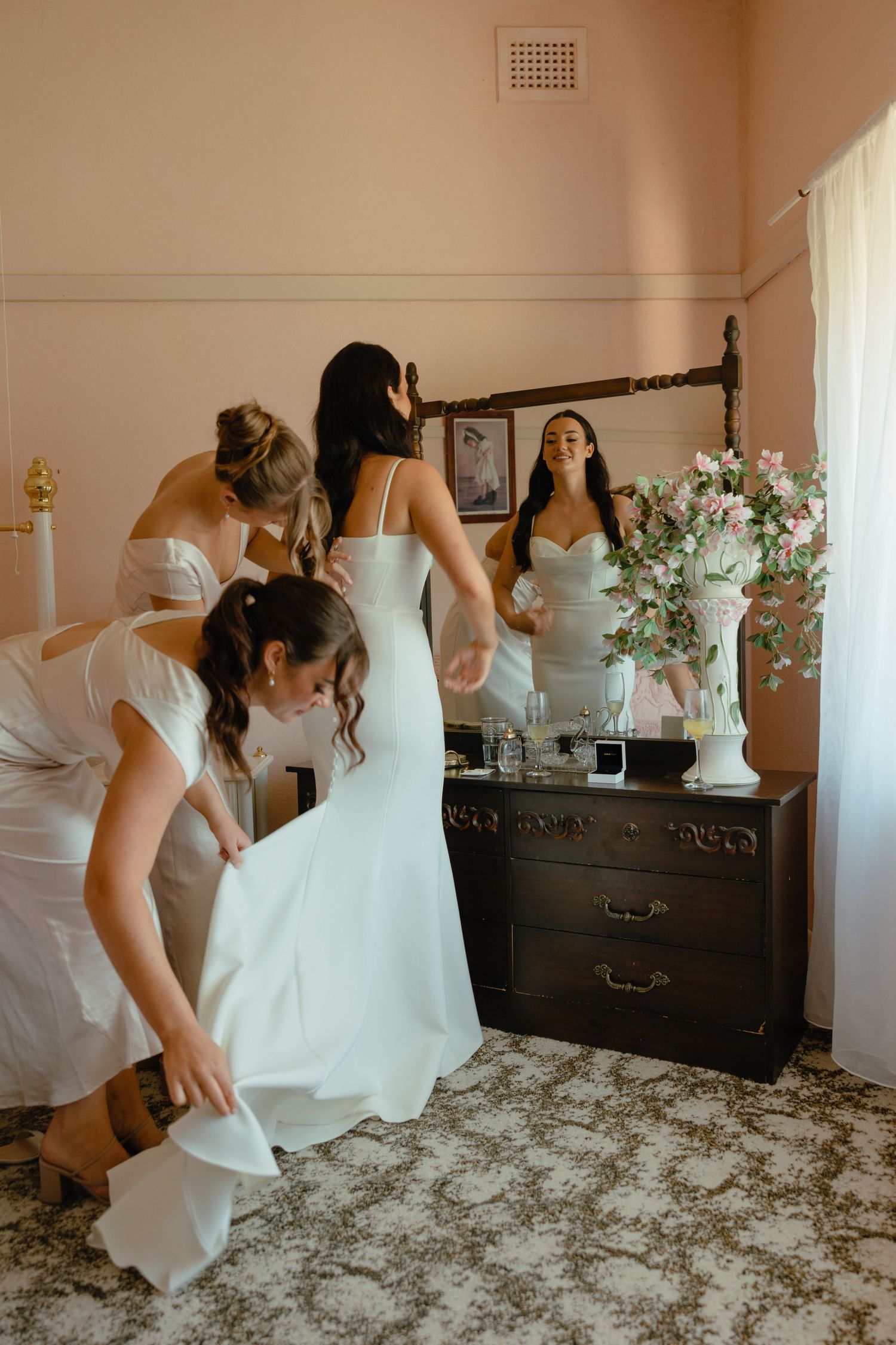 Wedding preparation scene with bridesmaids helping in a bedroom with vintage furniture.