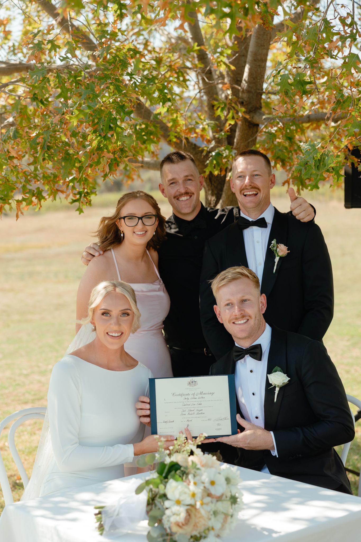 Wedding celebration group displaying marriage certificate under autumn tree branches on sunny day.