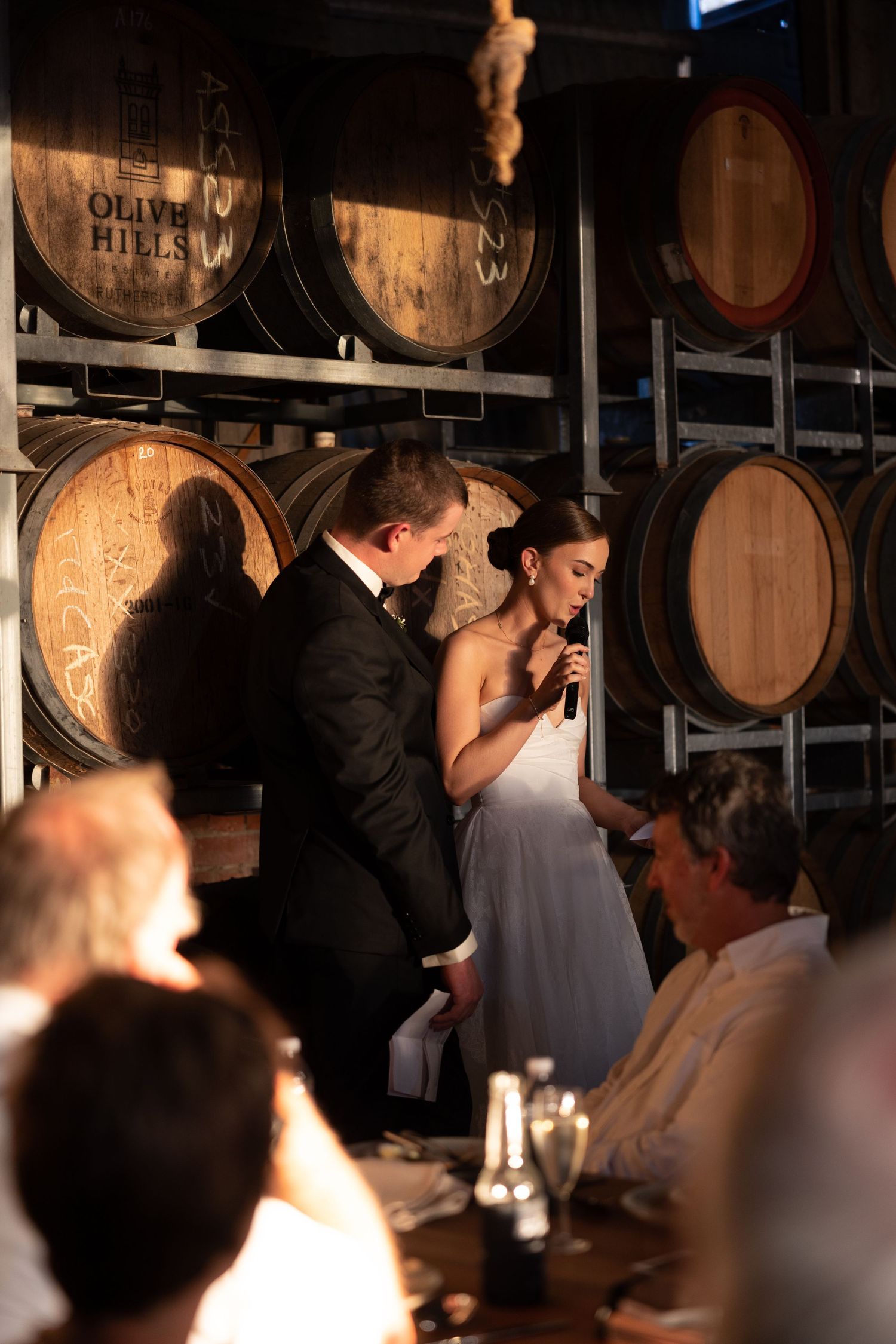 Wedding celebration in a rustic wine barrel room with guests watching speeches.
