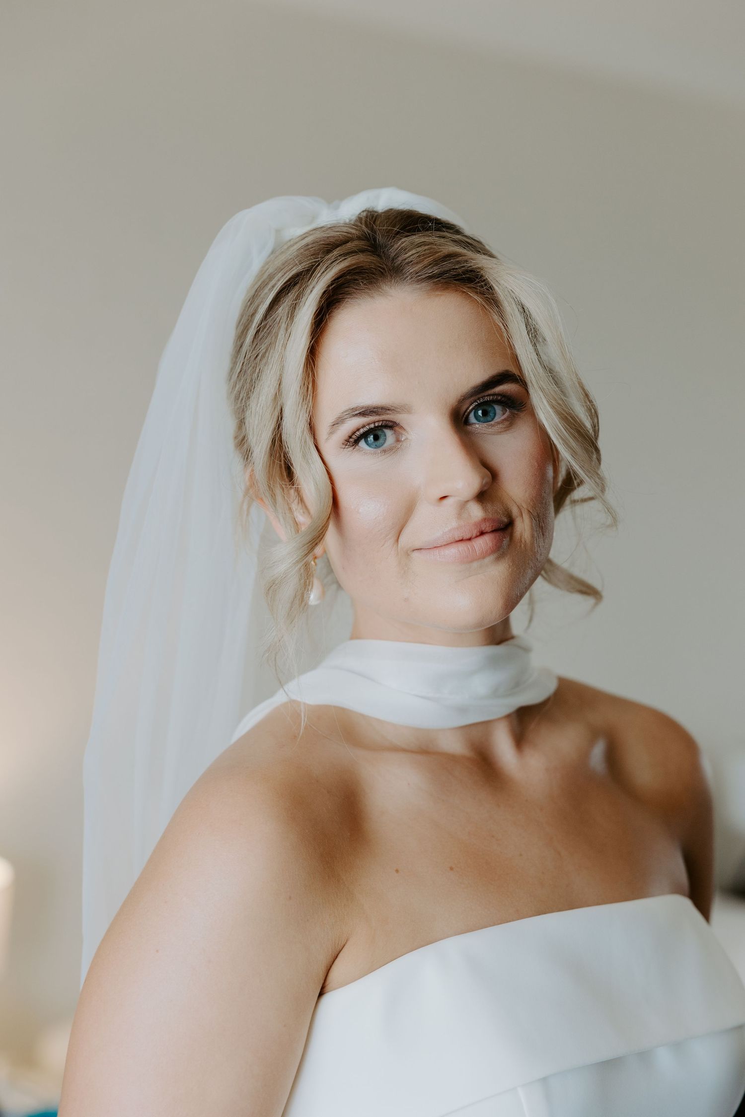 A bride wearing a white strapless wedding dress smiles softly while wearing a sheer veil against a neutral background.