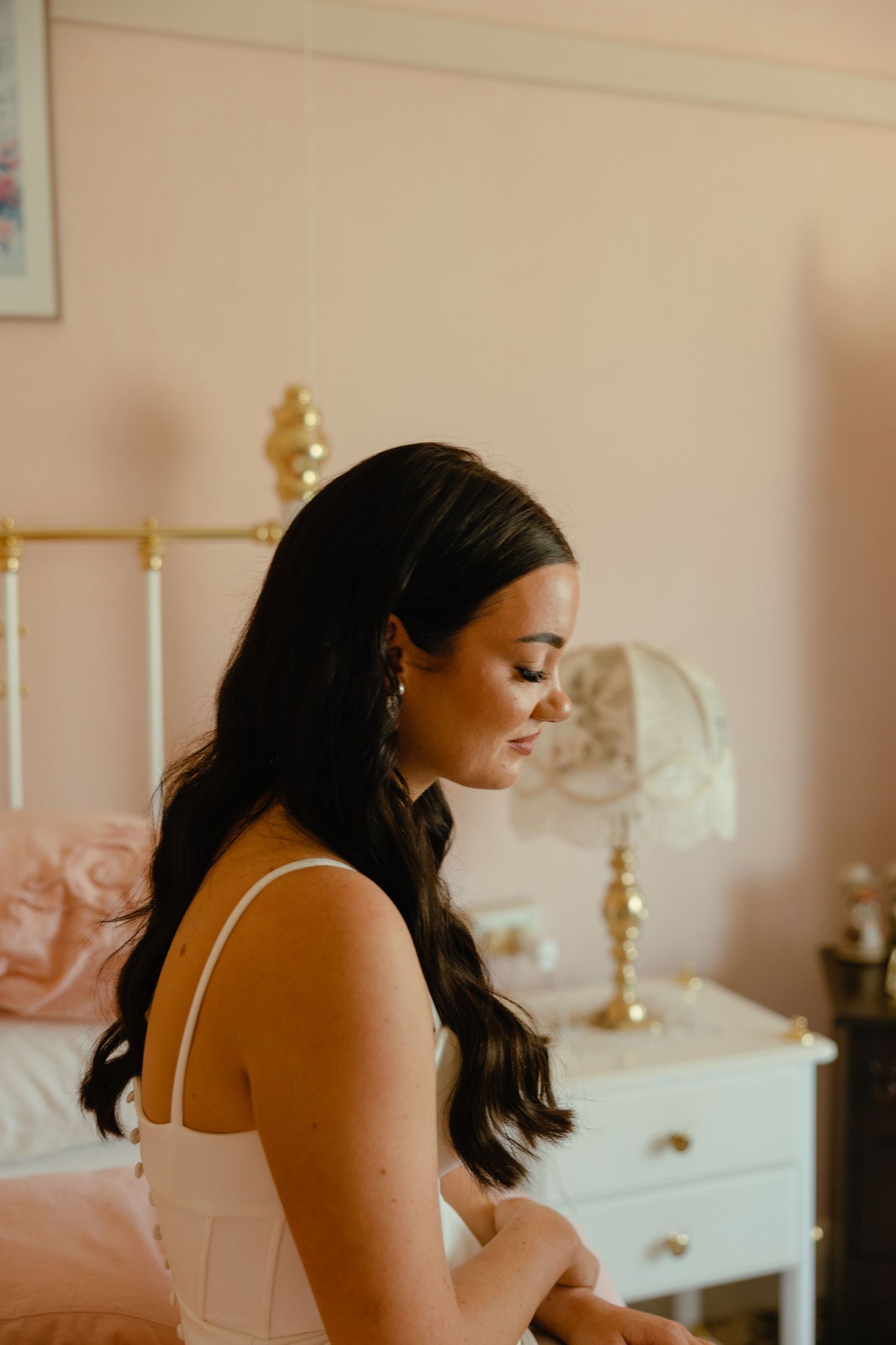 A bride in a white dress sits thoughtfully in a pink-toned bedroom setting.