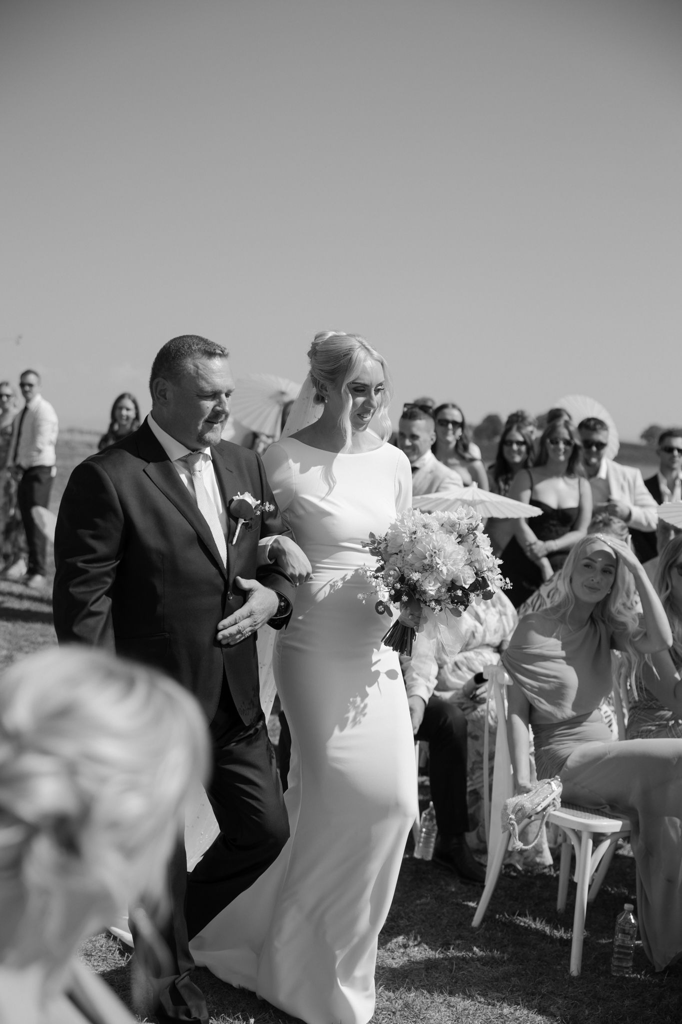 A couple walks down the outdoor ceremony aisle while wedding guests look on in black and white.