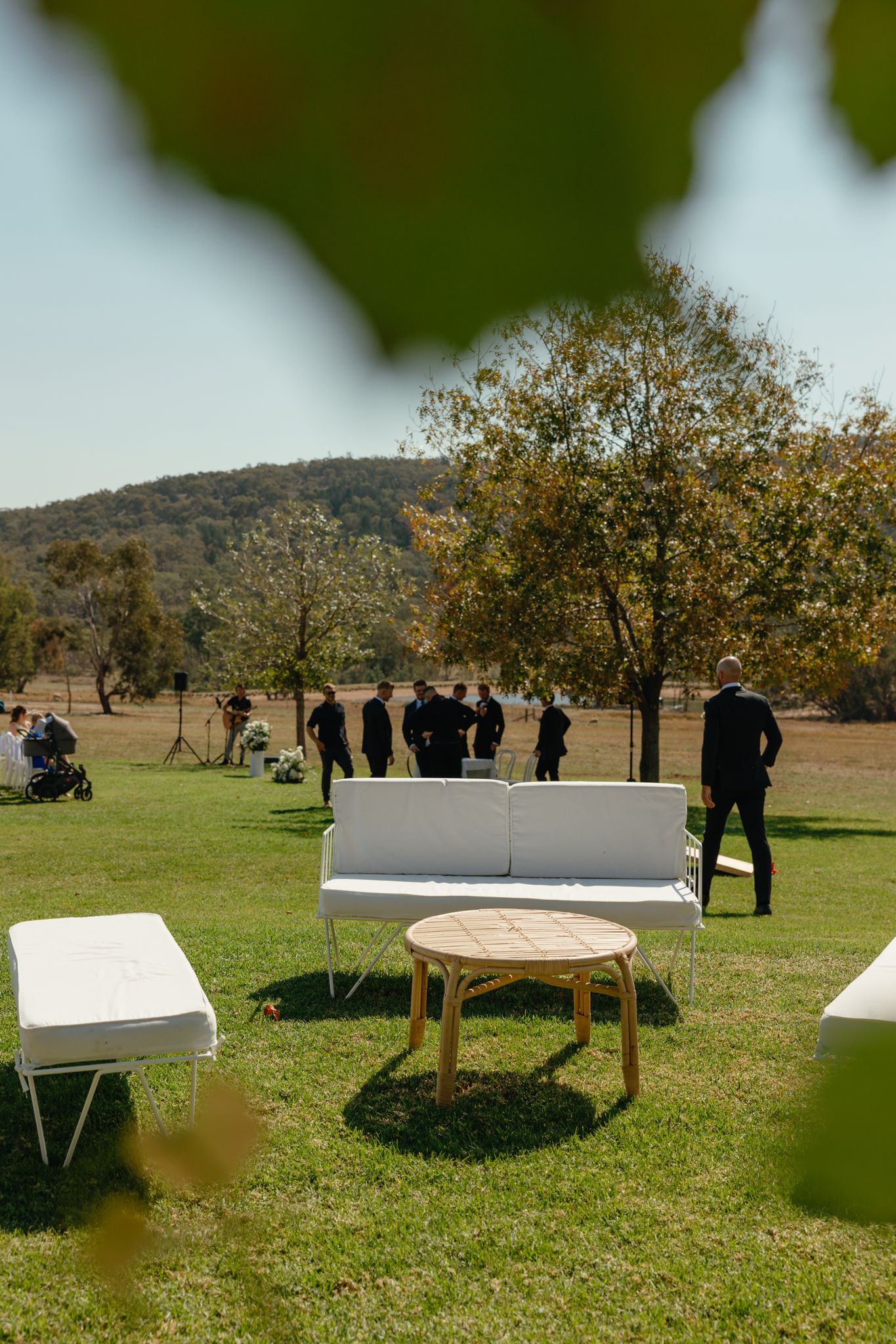White outdoor seating arrangement with a wooden table set up on grass lawn with hills and trees in the background.