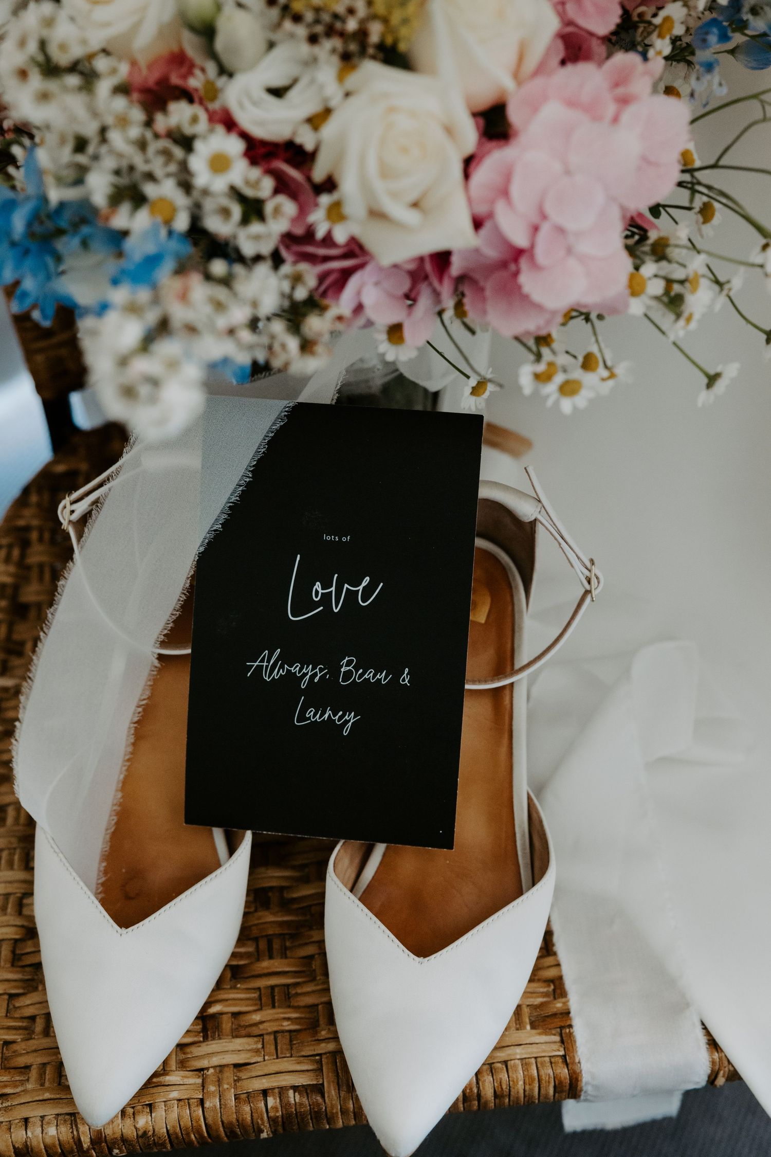 Elegant white wedding shoes with a black card reading 'Love' alongside pink and blue floral arrangements.
