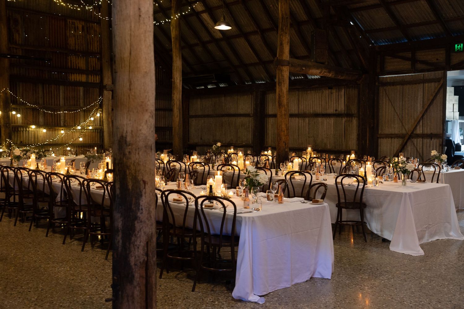 Rustic barn wedding reception venue with long tables, white linens, and twinkling string lights creating an elegant atmosphere.