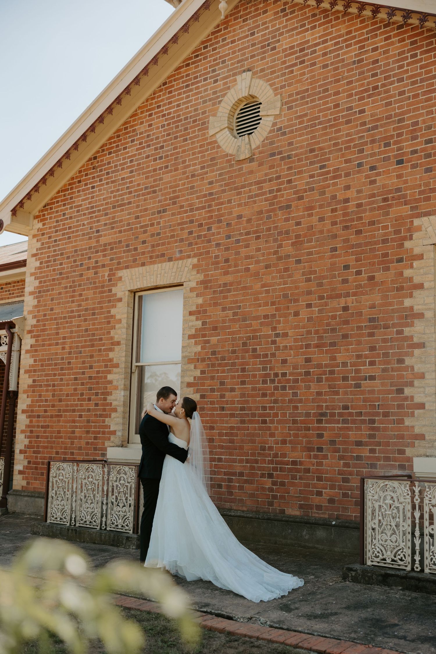A couple embraces in front of a rustic brick building with elegant architectural details and a decorative round window.