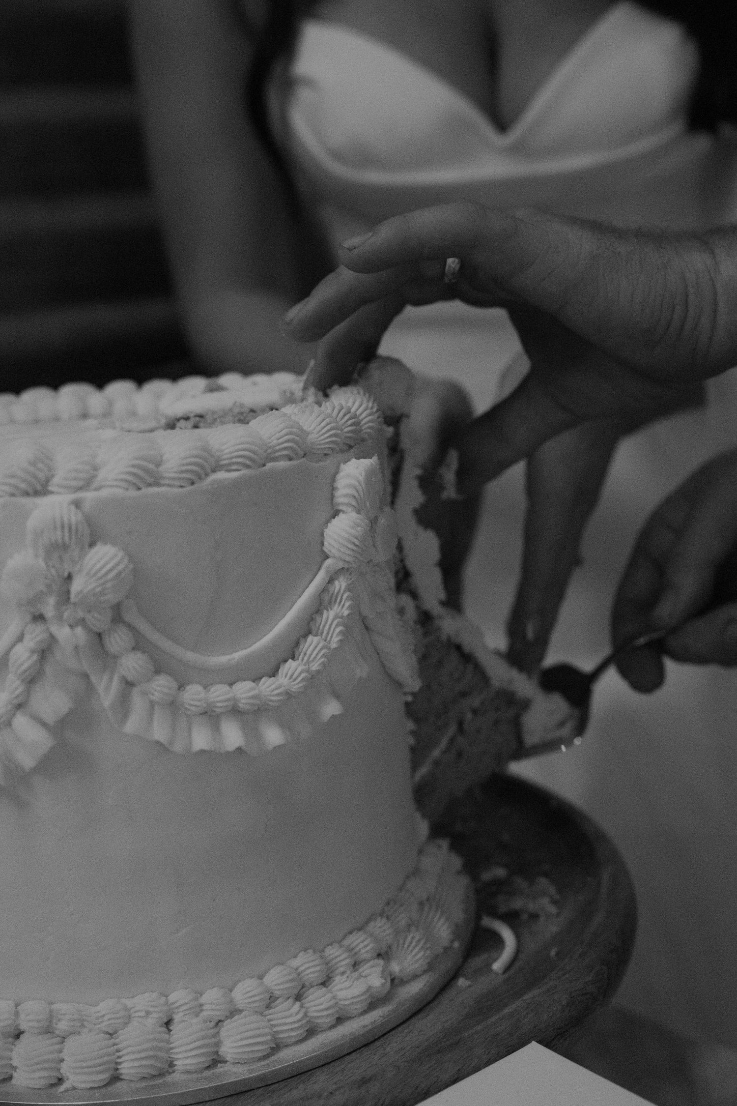 A close-up black and white photograph of hands decorating a wedding cake with delicate piped frosting details.