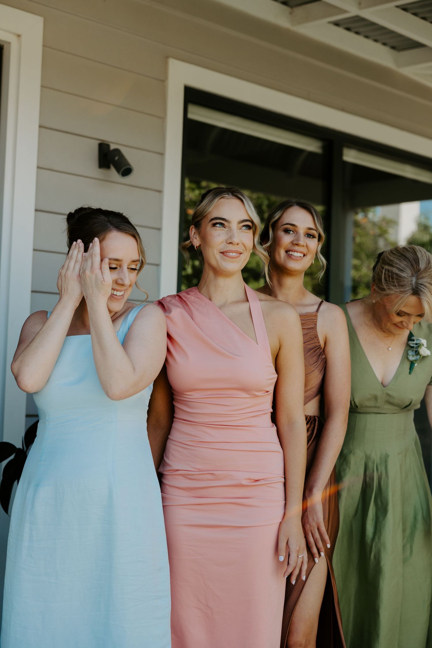 Bridesmaids wearing pastel dresses in blue, pink, and sage green pose together on a porch.