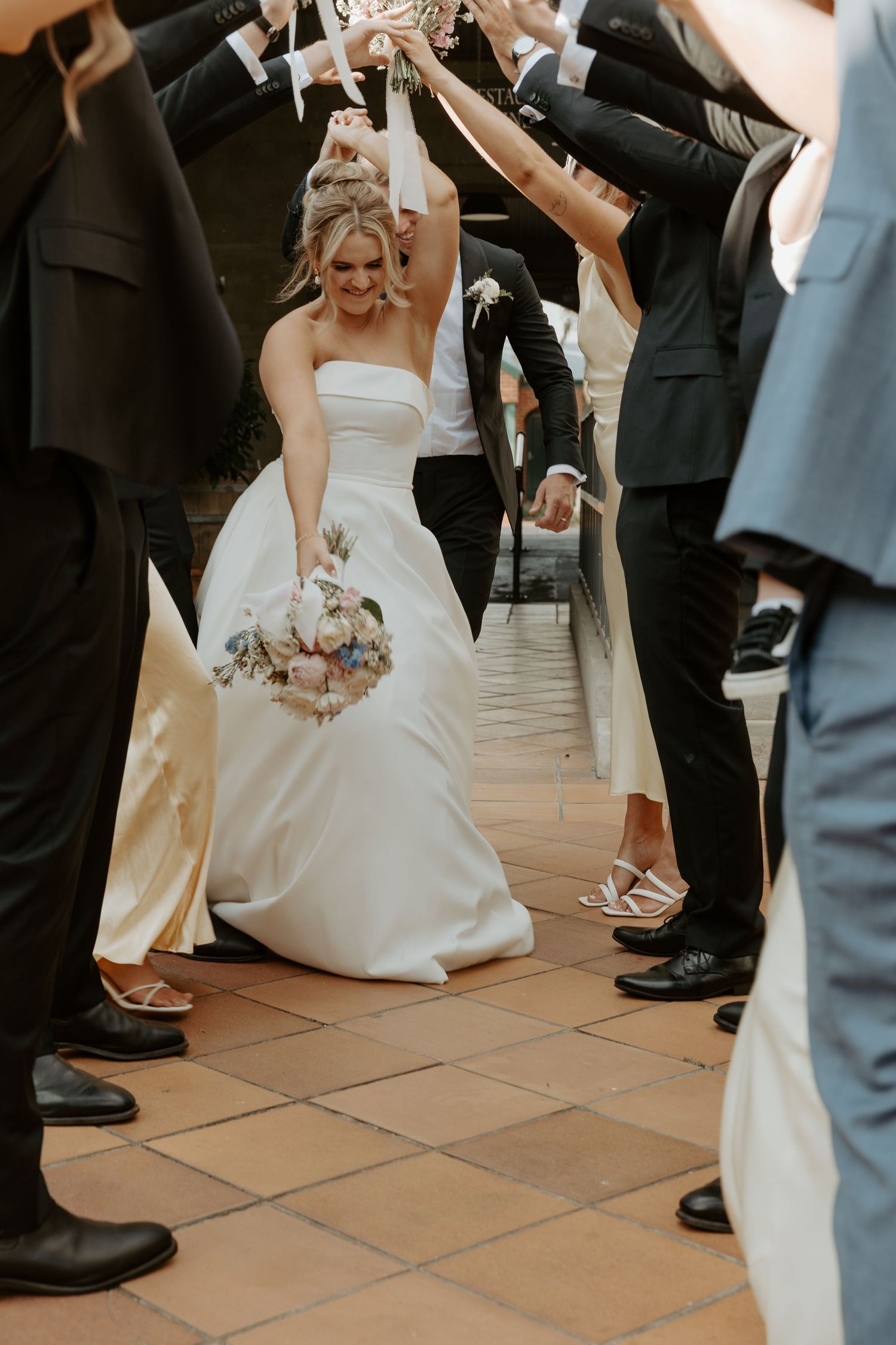 A bride in a white wedding dress holds a bouquet while passing under an arch of raised arms at her wedding ceremony.