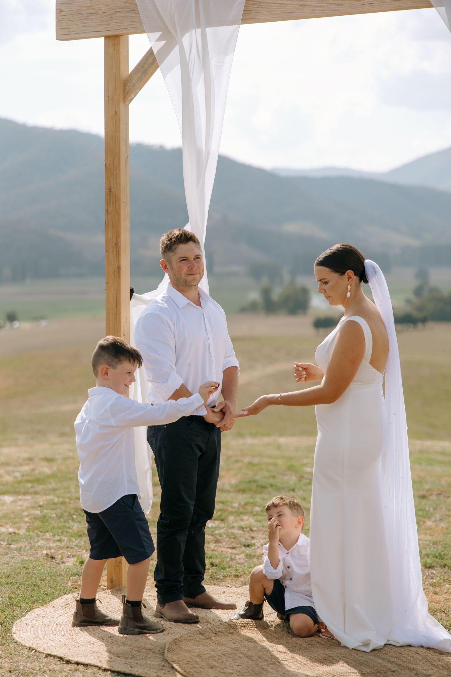 Intimate wedding ceremony moment under wooden arch with flowing white fabric and mountain views.