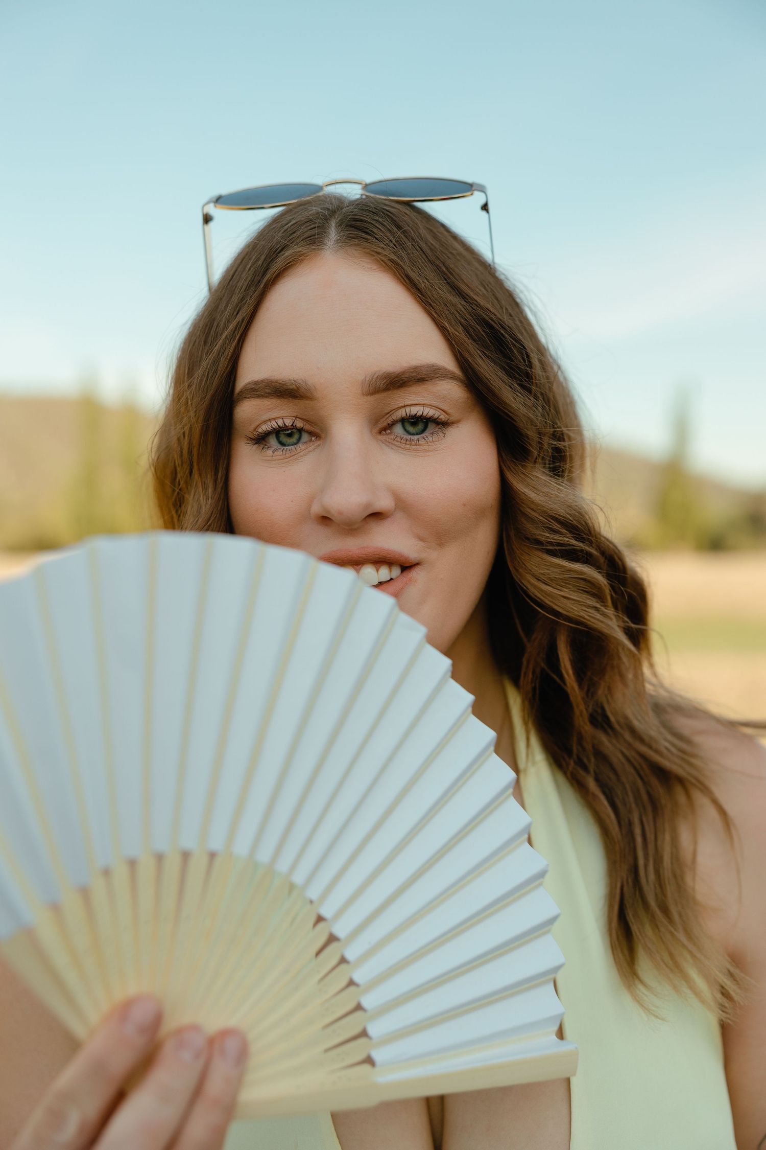 A folding paper fan held up against a sunny outdoor background with blue sky and sunlit scenery.