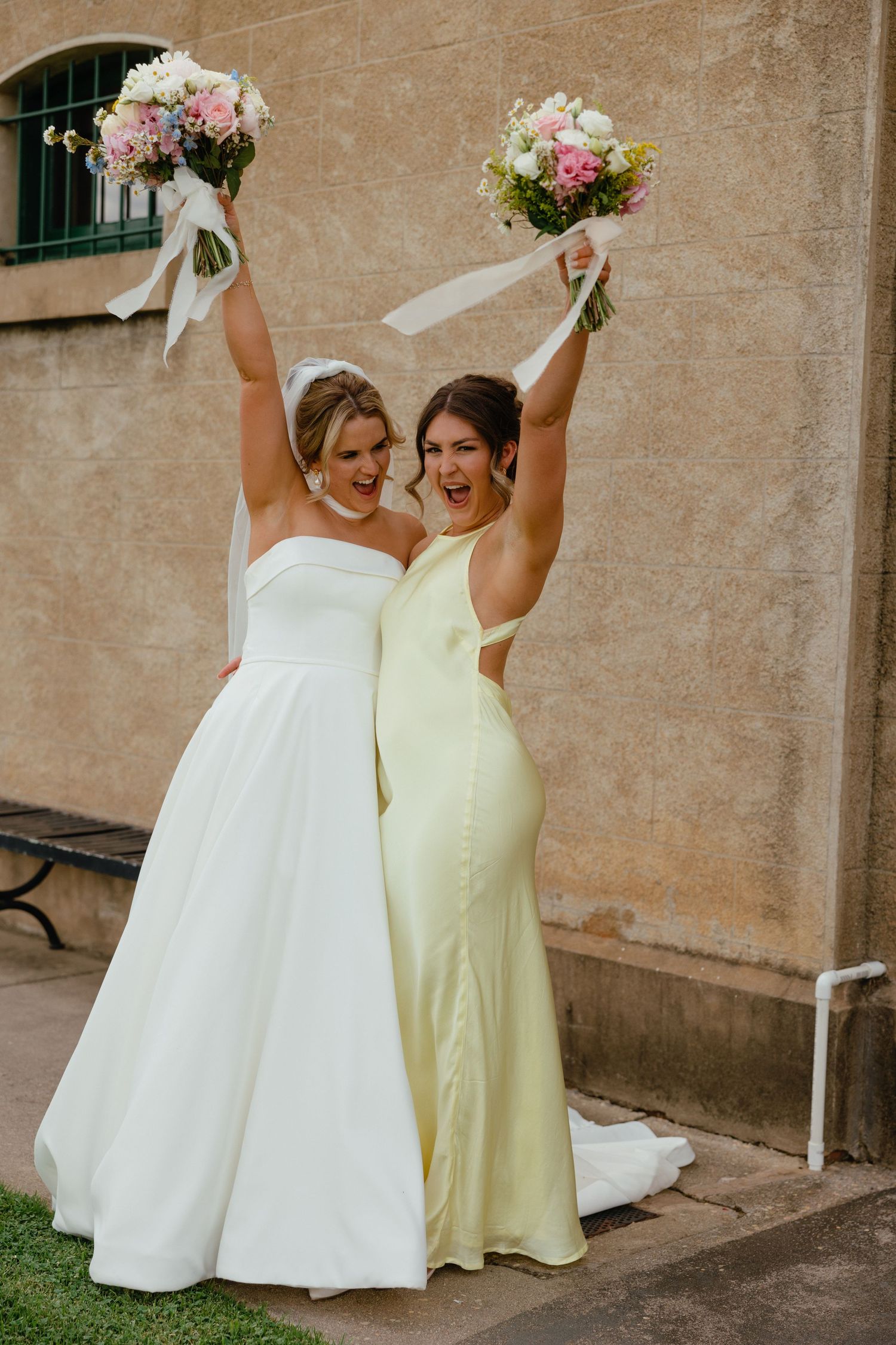 Two women in bridal and bridesmaid dresses joyfully hold up floral bouquets against a stucco wall.
