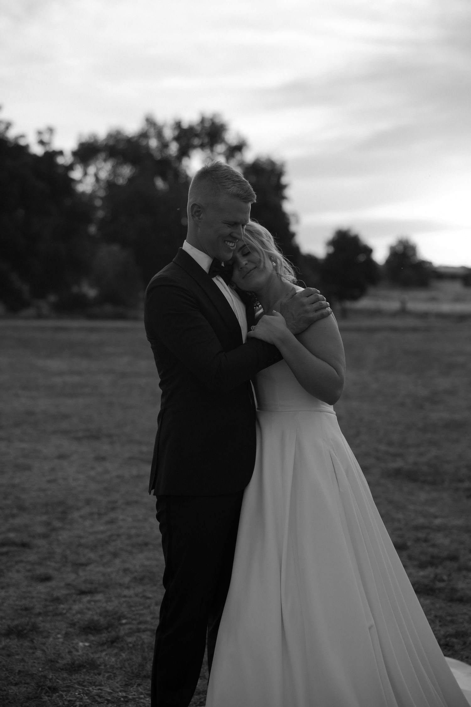 Black and white romantic portrait of couple in formal wedding attire embracing in outdoor setting.