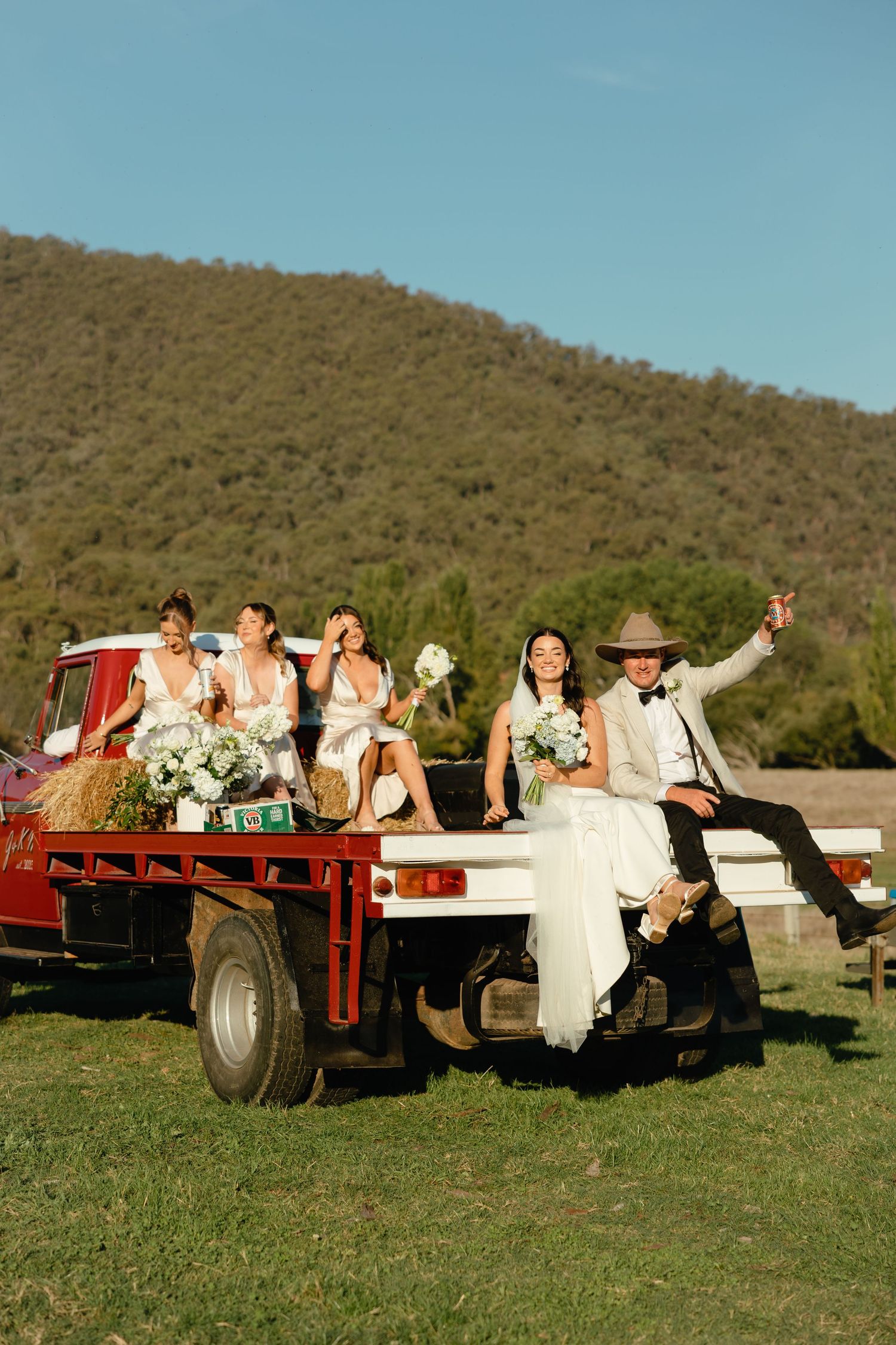 Wedding party riding on a rustic red farm trailer in front of a hill during a country outdoor celebration.