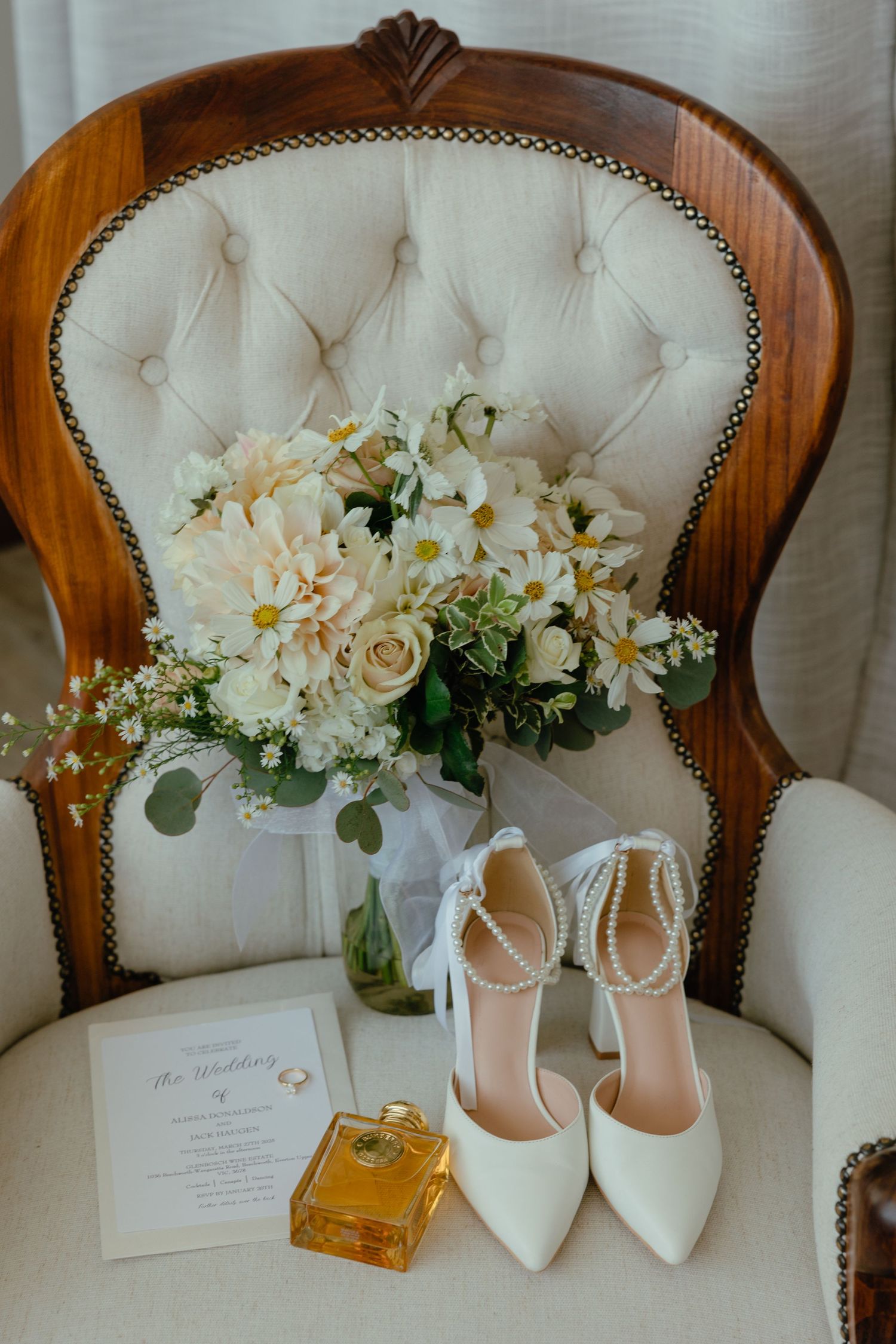 Elegant bridal details including white heels, perfume bottle, and blush floral bouquet on vintage tufted chair.