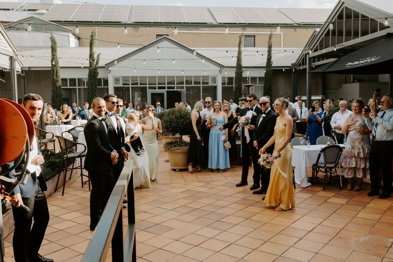 A large group of wedding guests gather on a tiled terrace for a celebration reception.