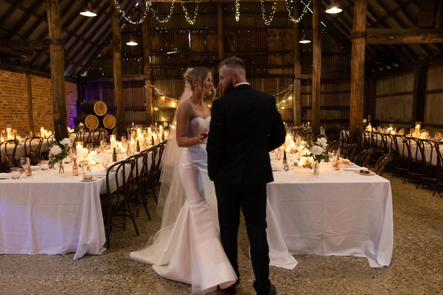A romantic wedding reception in a rustic barn venue with elegant white tablescapes and twinkling string lights overhead.