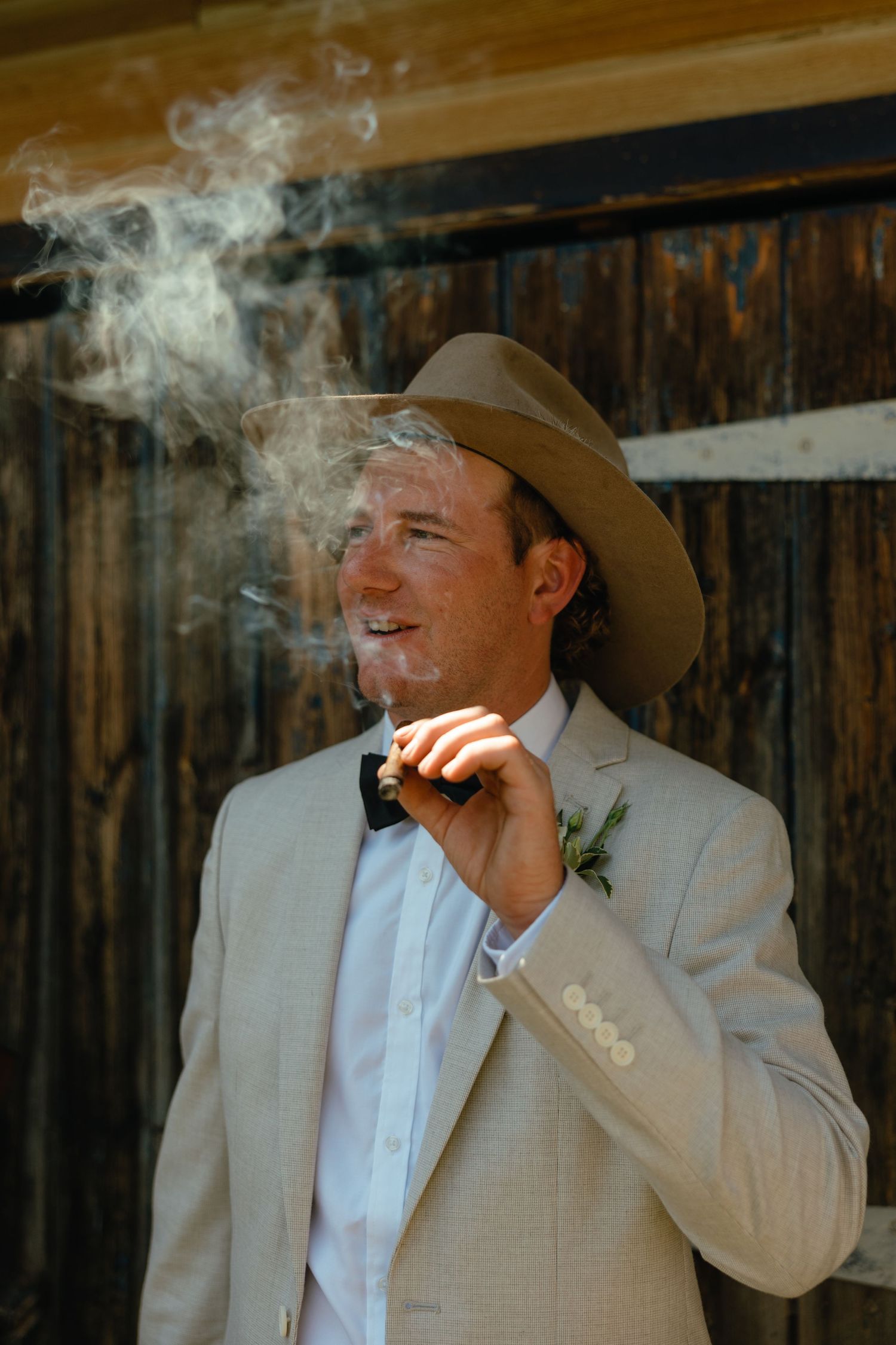 A groom in a light suit and cowboy hat smokes while standing against a rustic wooden wall.