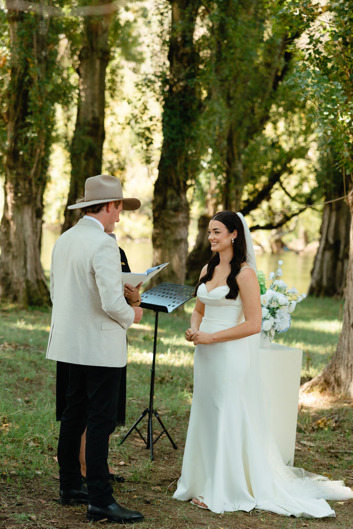 A wedding officiant in a hat and light jacket conducts an intimate outdoor ceremony beneath towering trees.