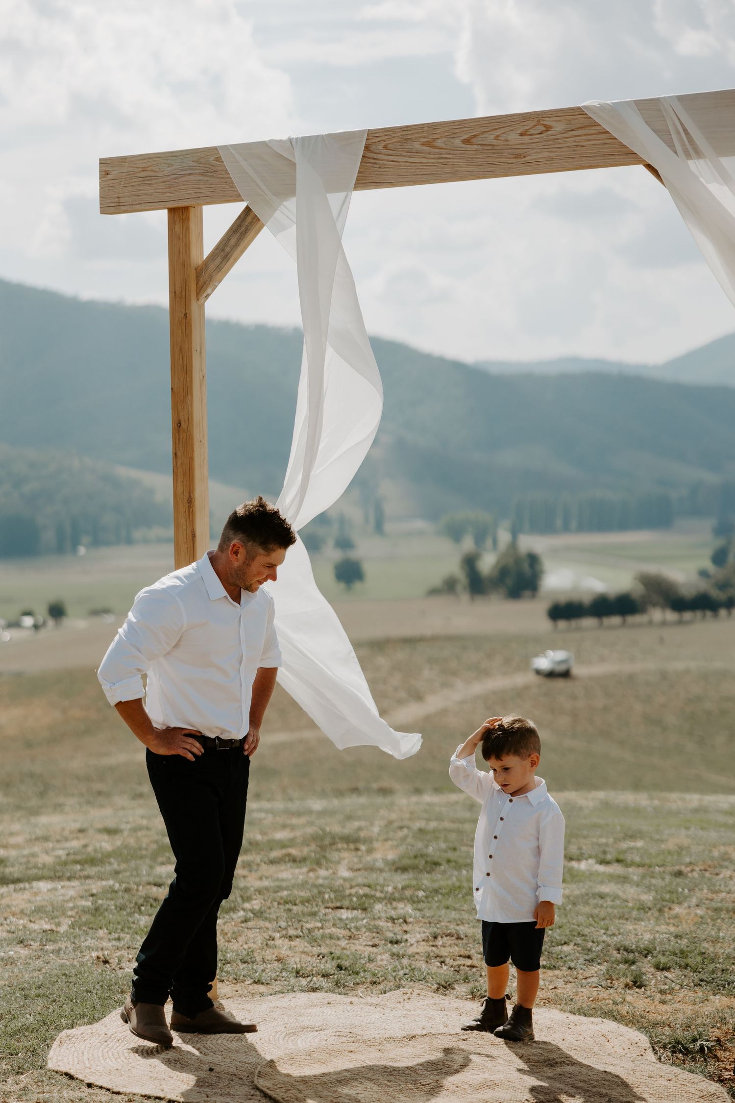 Man in white shirt looking down at small boy under wedding arch in mountain setting.