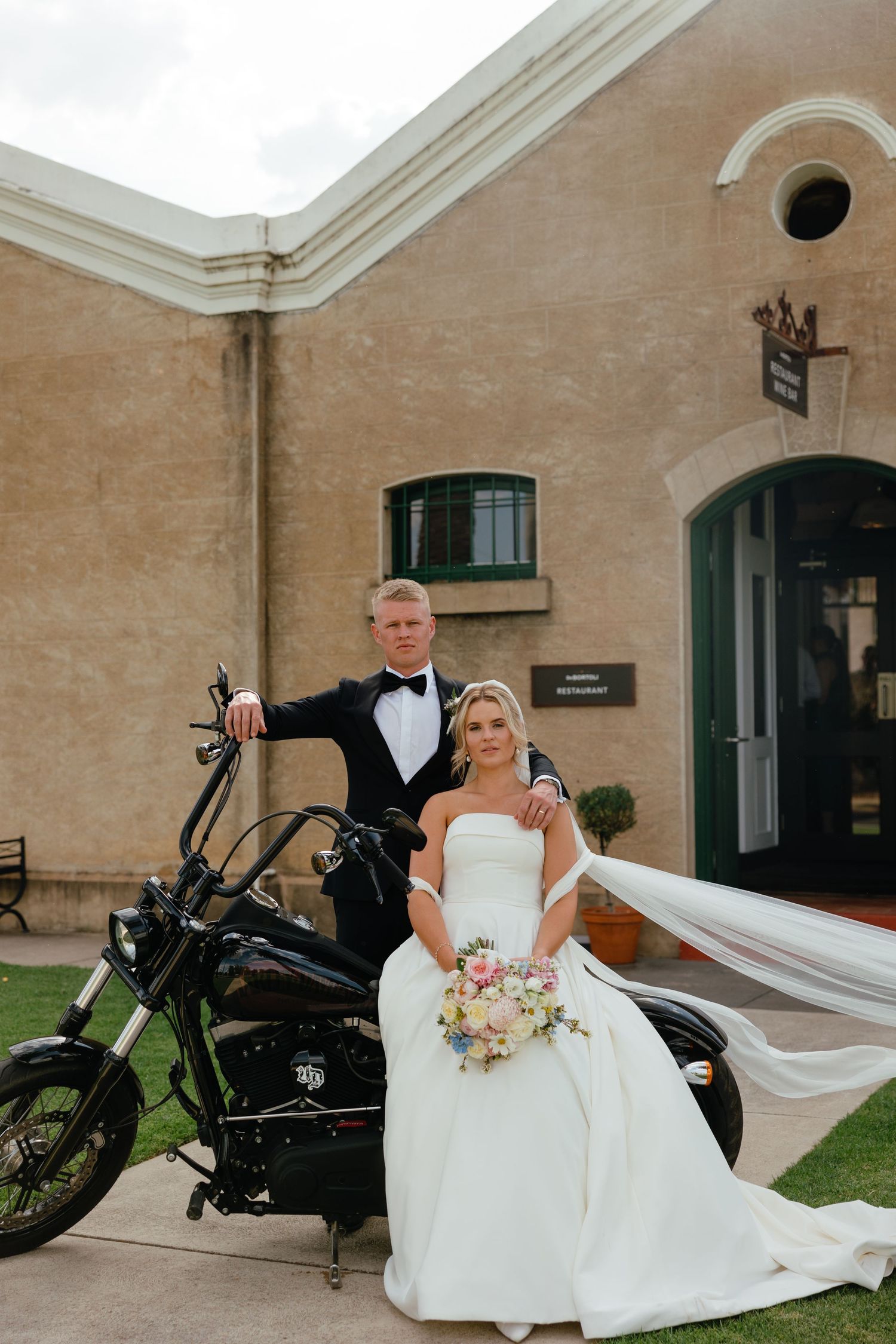 Wedding couple poses with vintage motorcycle outside stone building with elegant formal attire.