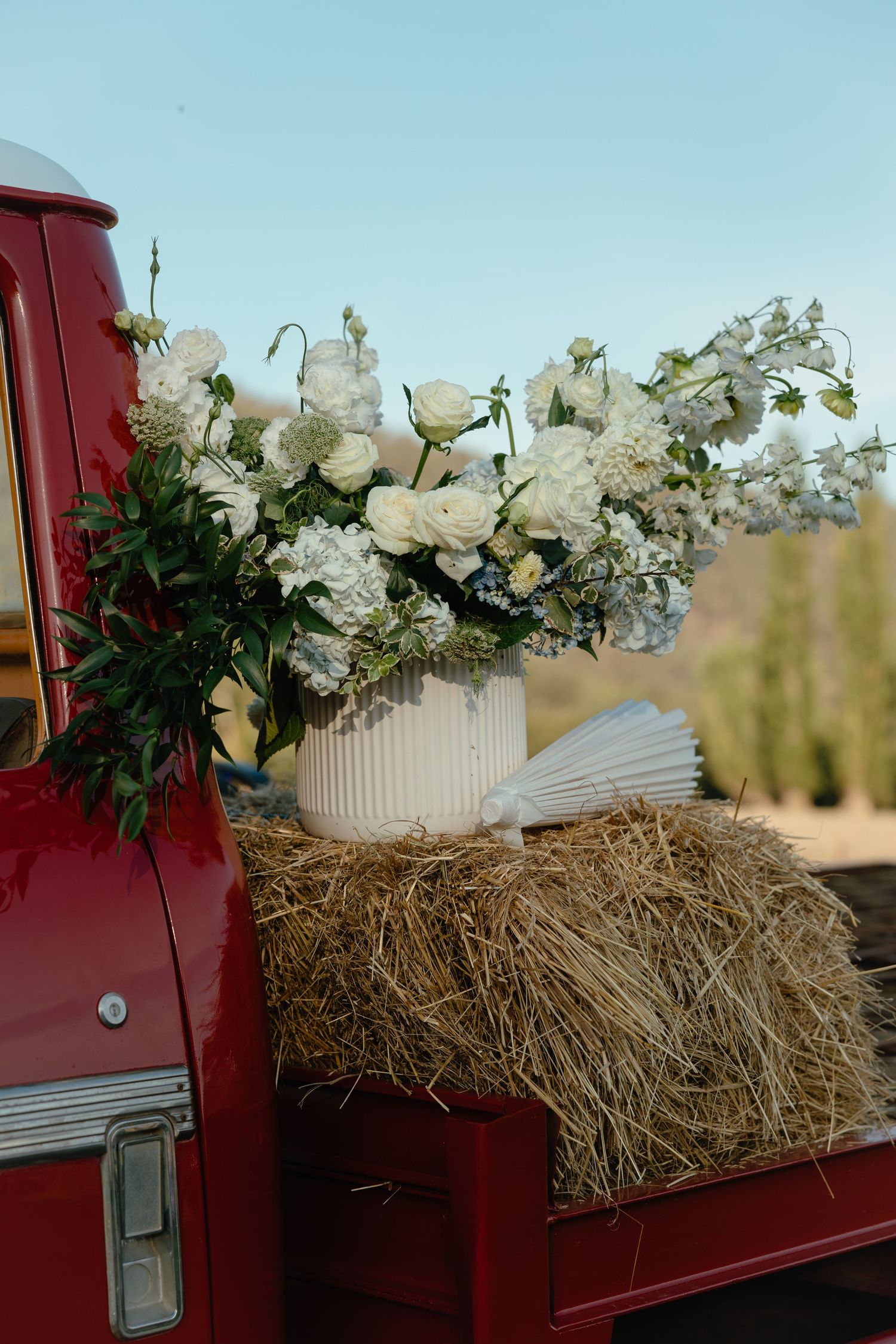White roses and floral arrangements displayed in a ribbed vase on hay bales in the bed of a vintage red pickup truck.