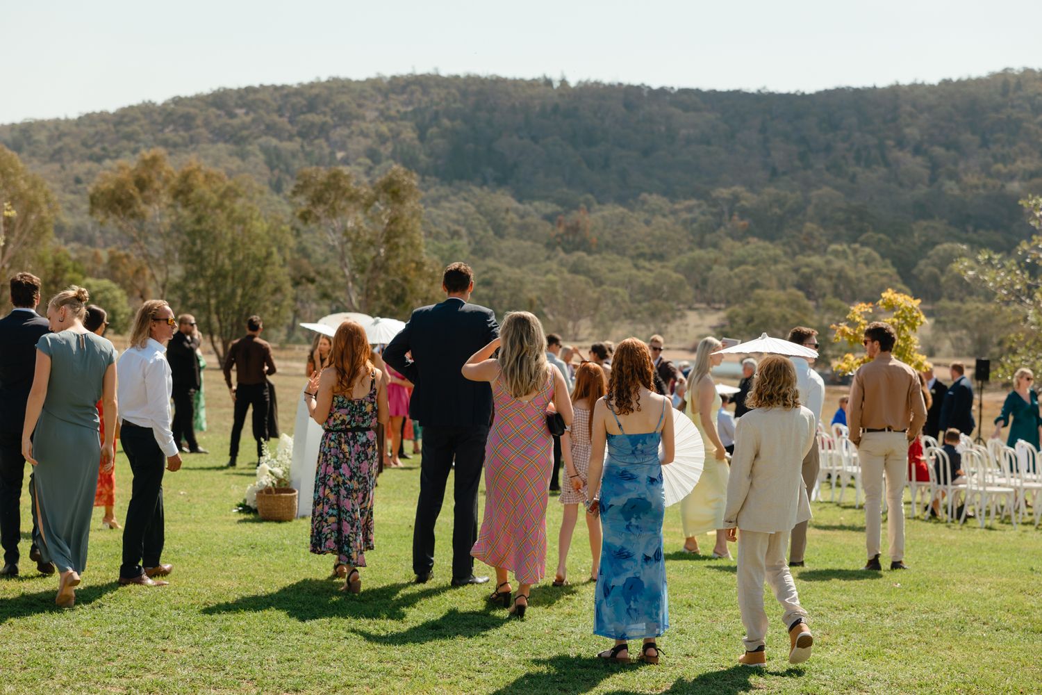 Wedding guests walk across a sunny meadow surrounded by rolling hills during an outdoor celebration.
