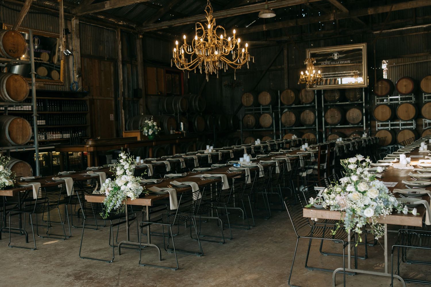Rustic winery reception venue with elegant chandeliers, wine barrels, and white floral centerpieces on long tables.