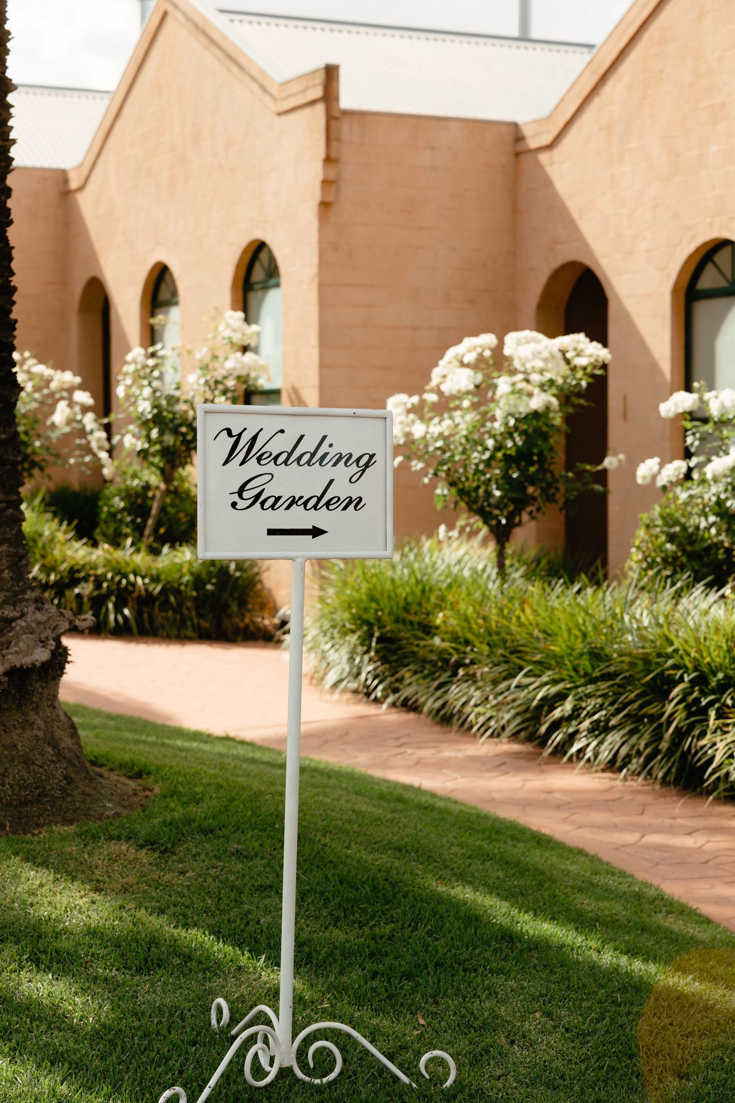 A white decorative sign reading 'Wedding Garden' stands in front of a beige building with arched windows and white flowering plants.