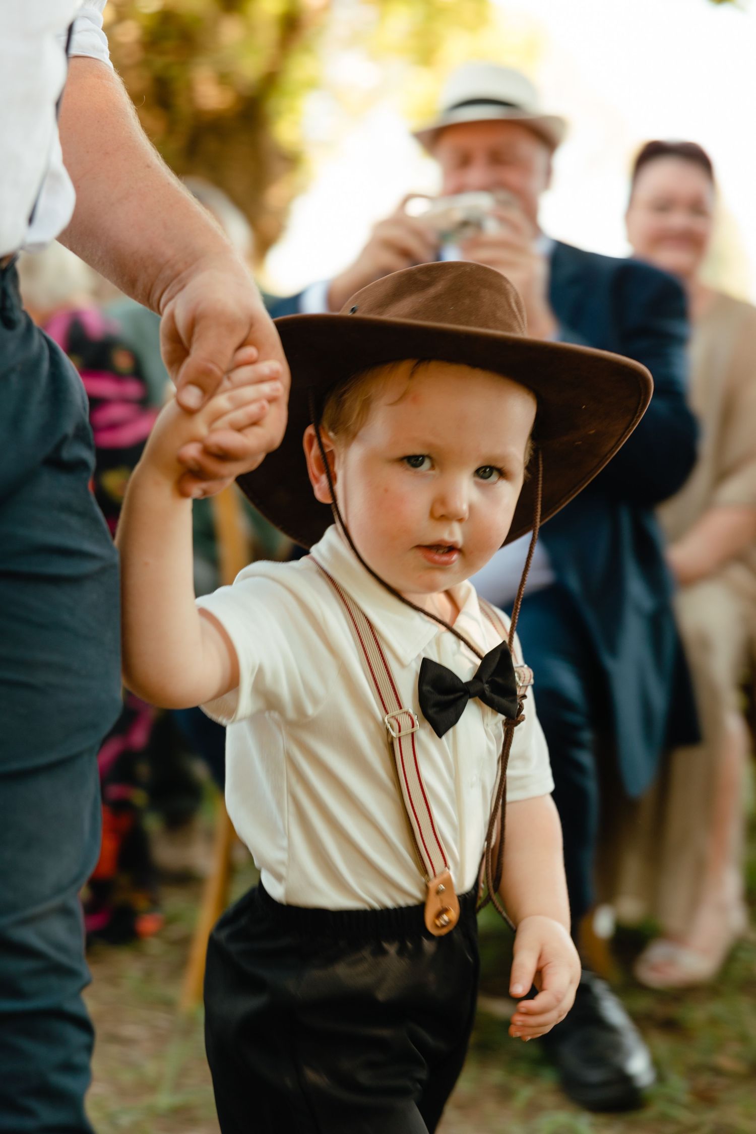 Small child dressed in formal vintage style with brown hat and bow tie holds an adult's hand at an outdoor event.