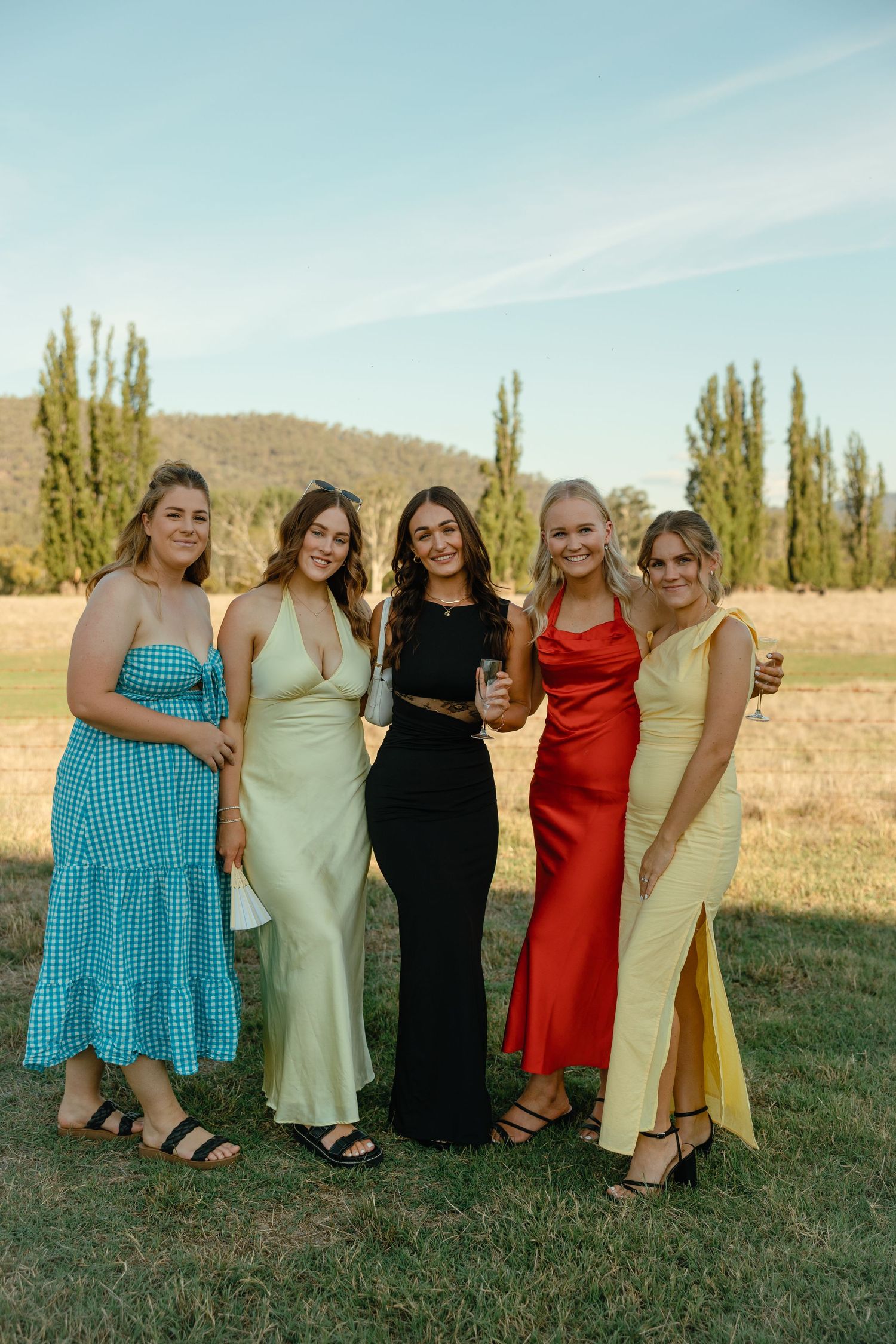 A group of five women in elegant formal dresses stand together in a field with tall poplar trees and hills in the background.