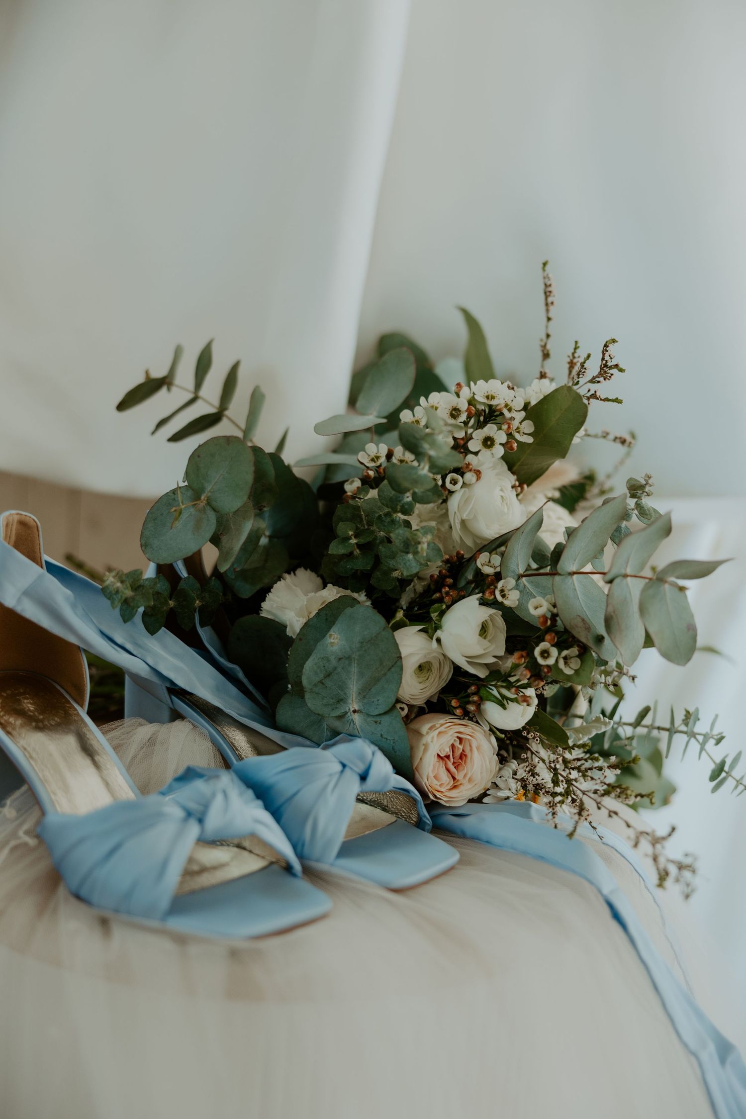 Delicate wedding details featuring blue bow sandals and eucalyptus floral arrangement.