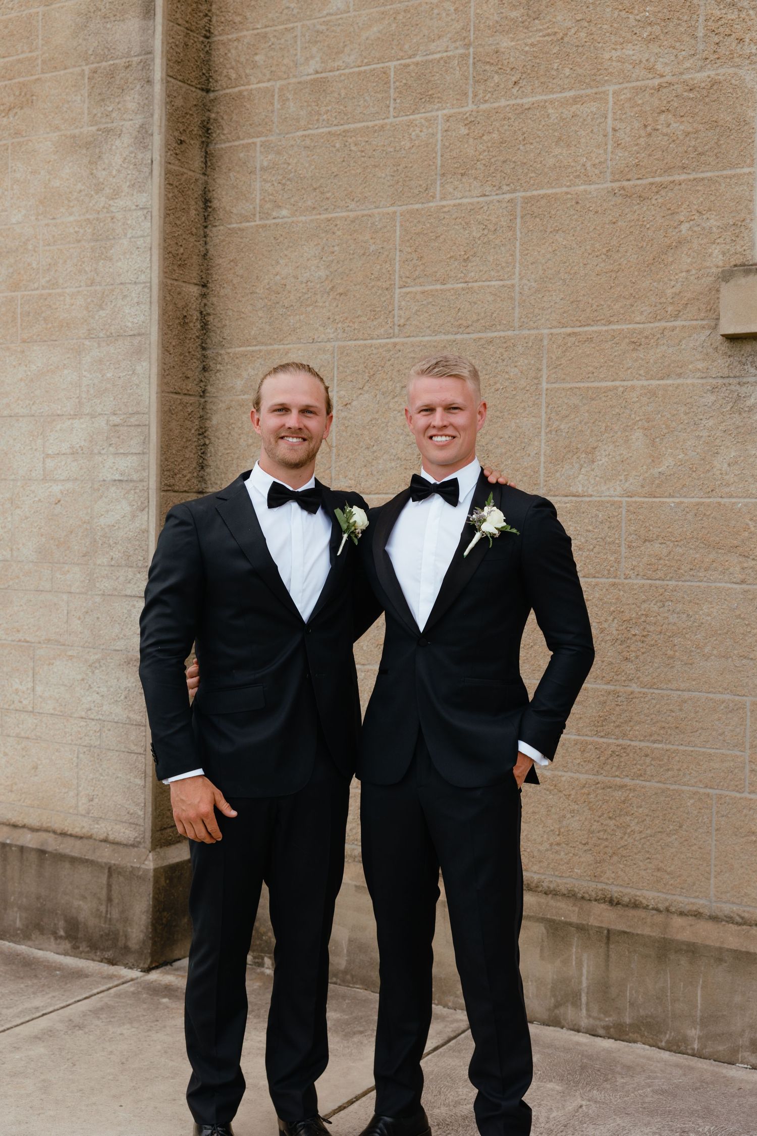 Two dapper groomsmen in classic black tuxedos and bow ties stand smiling against a stone wall at a formal wedding.