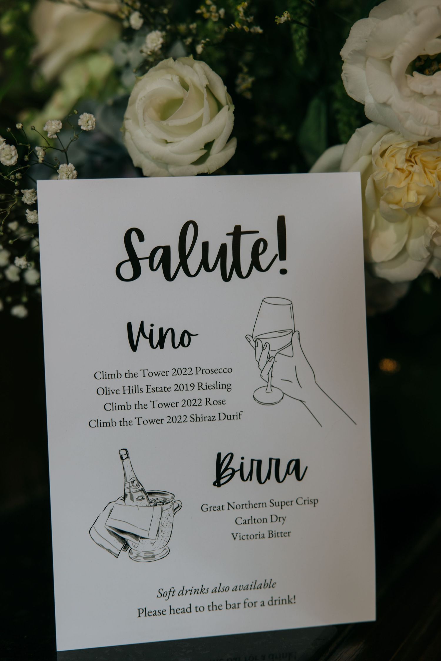 Wedding drink menu card featuring wine and birra selections displayed among white roses.