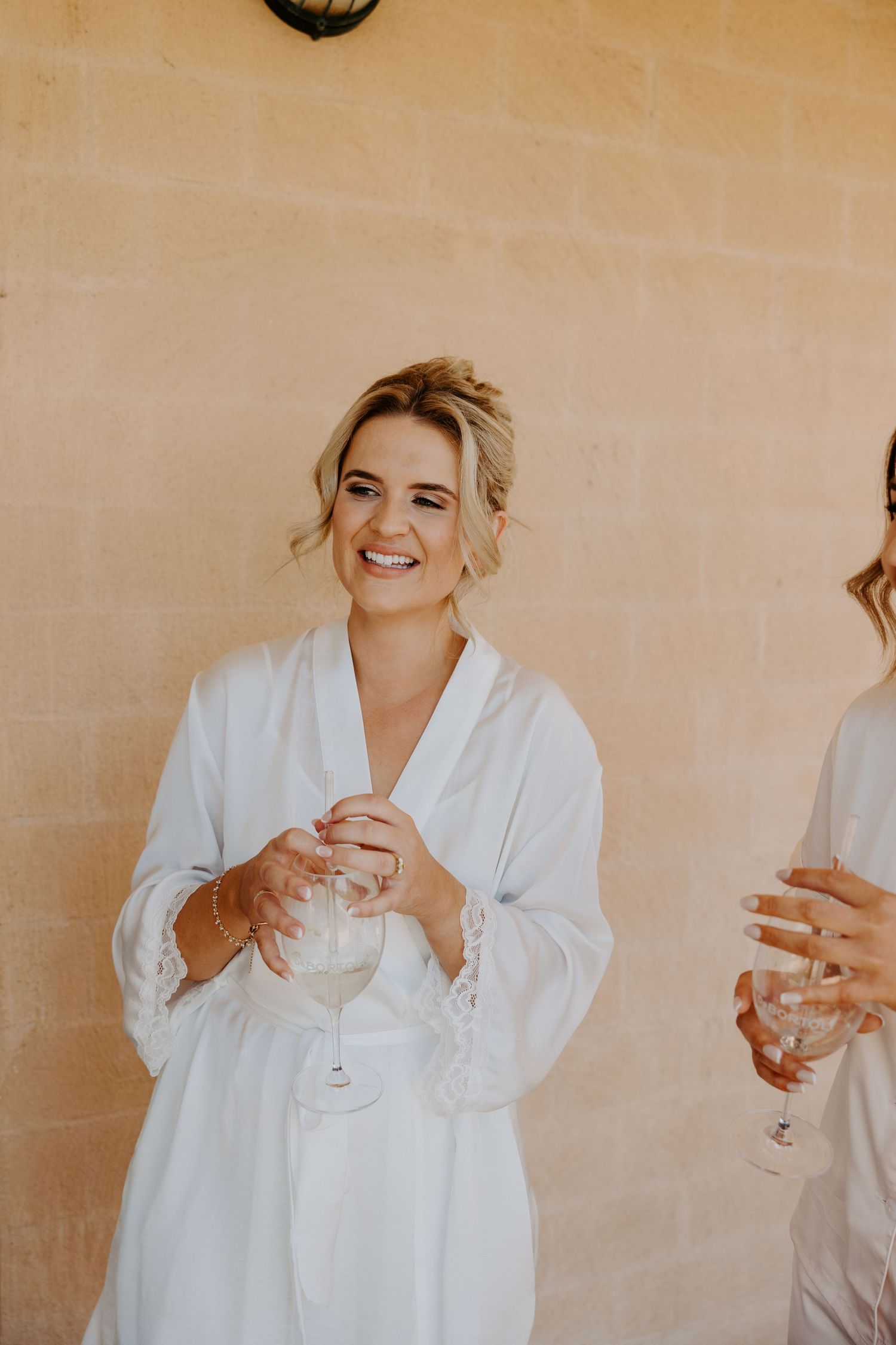 A person in a white robe holding a champagne glass smiles against a beige wall backdrop.
