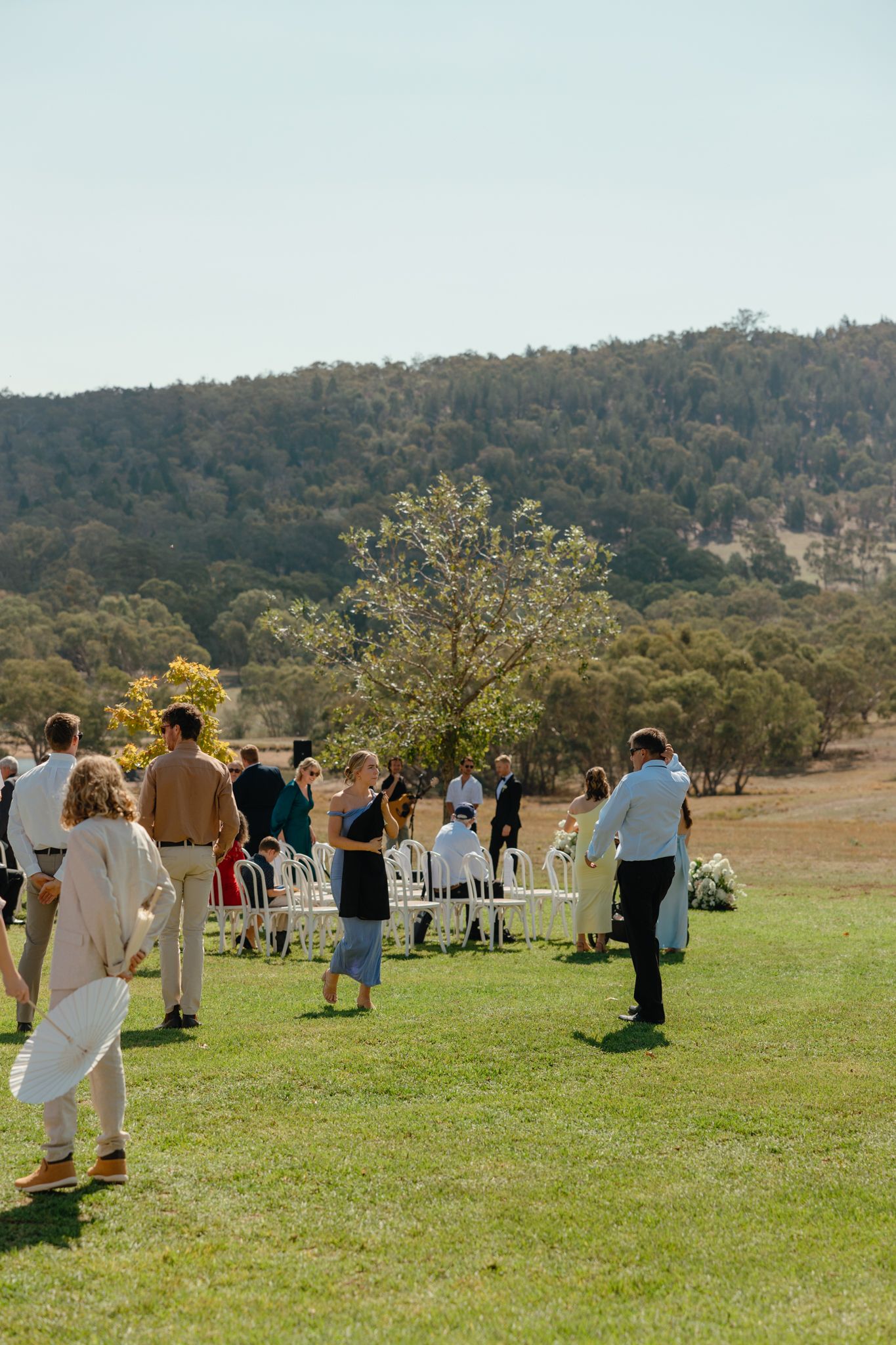Guests gathering at an outdoor wedding ceremony setup with white chairs on a grassy hilltop surrounded by mountains.