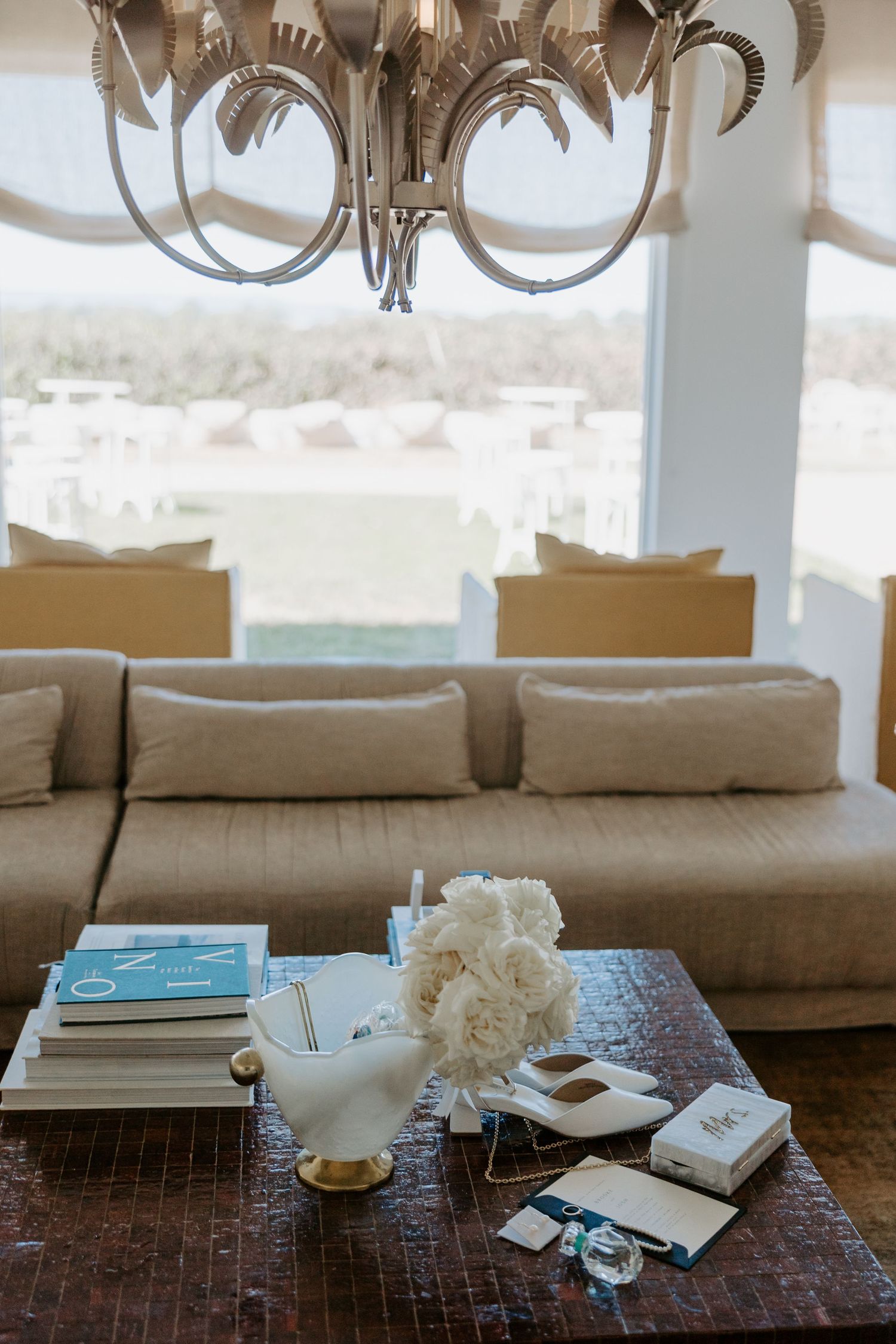 Elegant living room with beige sectional sofa, chandelier, coffee table display of books and white flowers against snowy view.