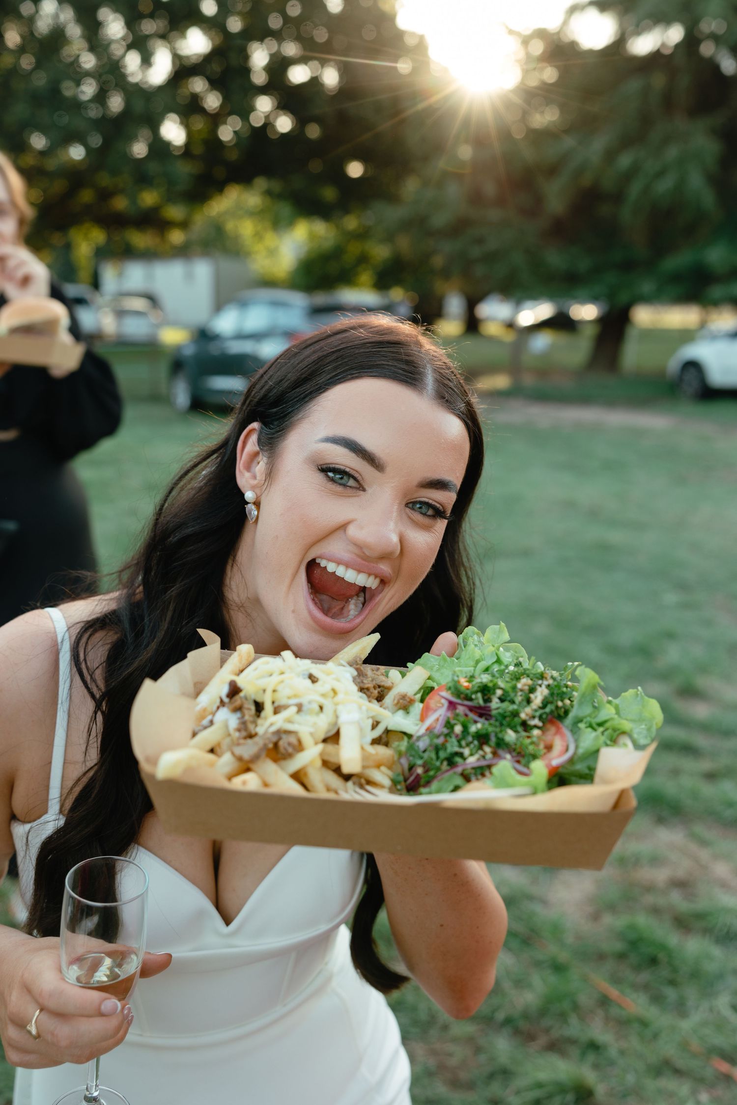 Woman in white dress holds a tray of fresh salad while laughing outdoors at sunset with bokeh background.