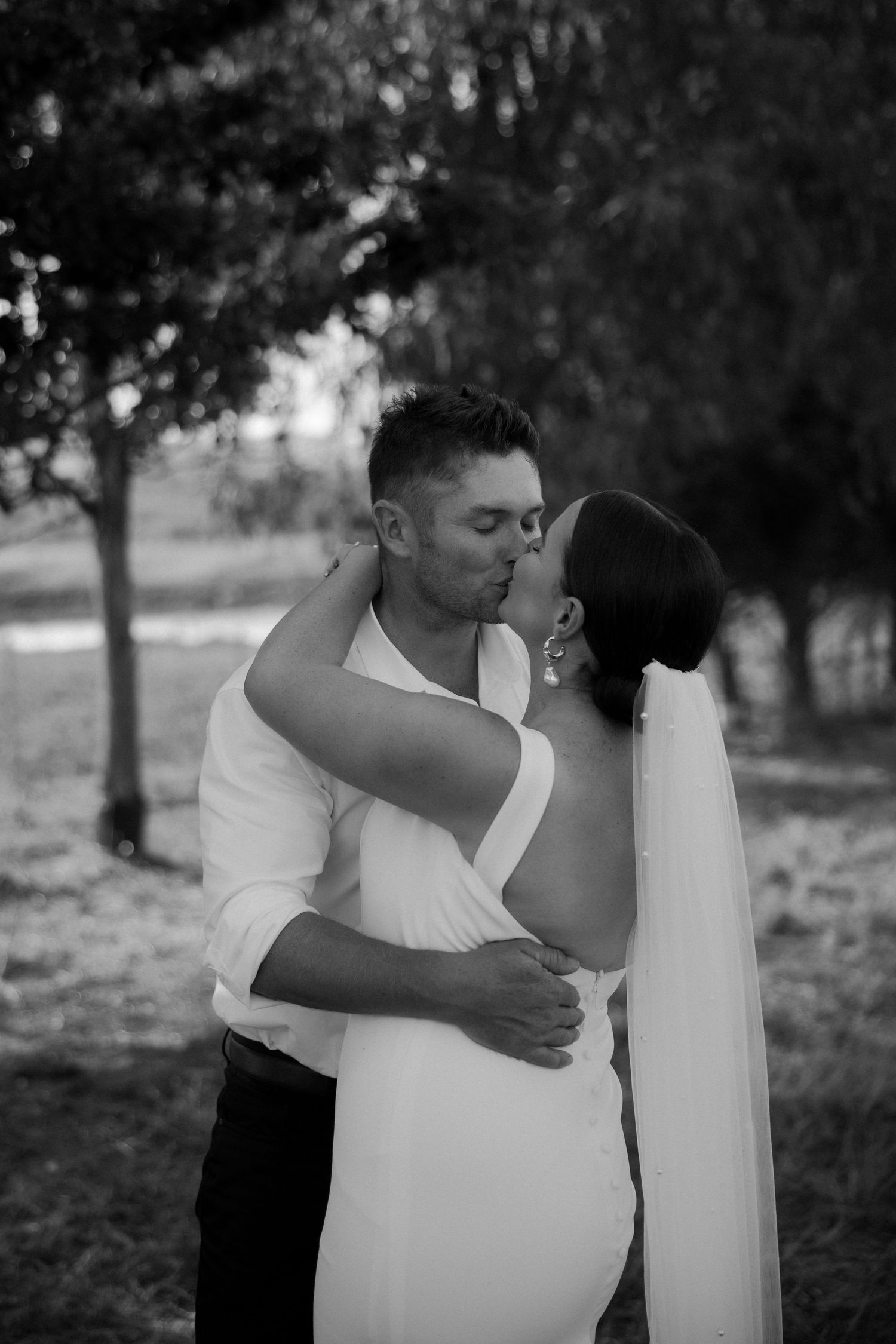 A romantic black and white wedding photo of a couple embracing outdoors under trees.