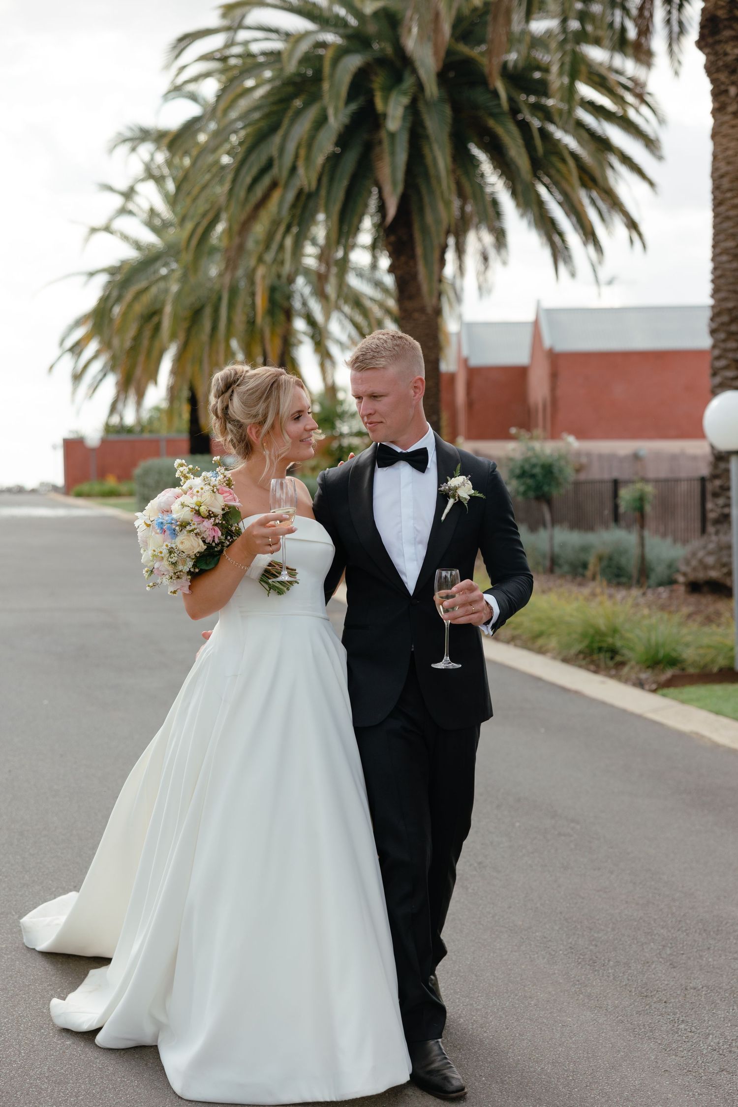 A newlywed couple walks together on a palm-lined pathway, the bride in white gown holding flowers and groom in tuxedo with wine.
