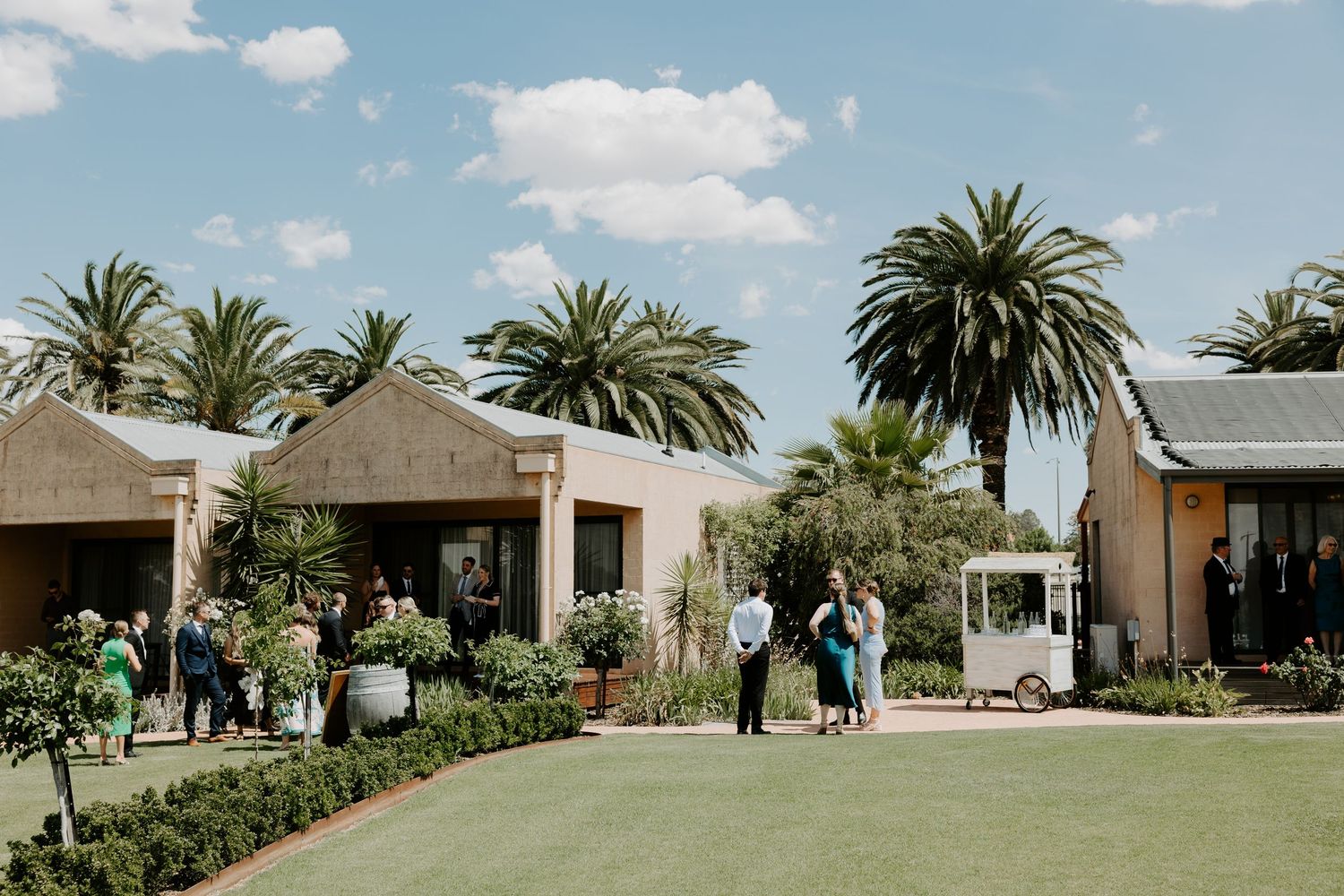 Outdoor wedding venue with palm trees and thatched roof buildings surrounding a manicured lawn on a sunny day.