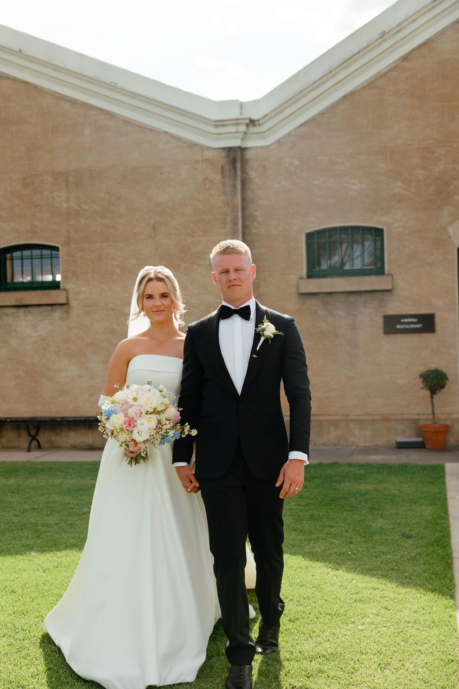 Wedding couple poses together on green lawn outside tan building with small windows.